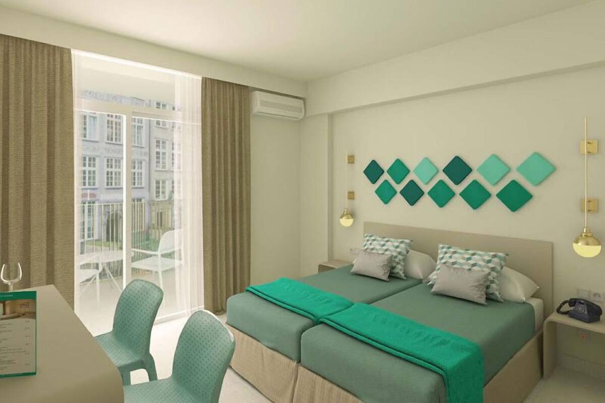 A hotel room with a bed, two bedside tables, a desk, and a chair. There is a window with curtains, and a wall-mounted air conditioning unit.