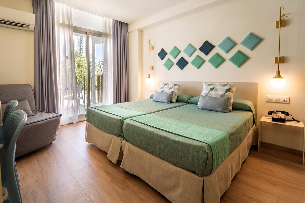 A hotel room with a double bed, a chair, a bedside table with a phone, and a view of the outdoors through a large window.