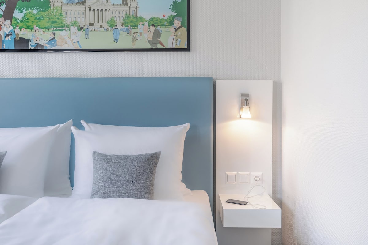 A neatly made bed with white and gray pillows, a blue headboard, and a bedside table with a lamp and a phone.