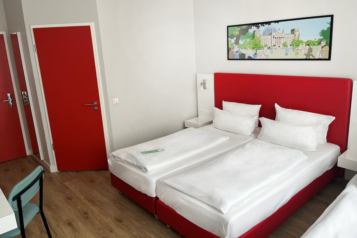 Hotellihuone, jossa on kaksi erillistä sänkyä, punainen pääty, valkoiset vuodevaatteet, vihreä kyltti yhdessä sängyssä, valkoinen yöpöytä, valkoinen työpöytä ja punainen ovi.