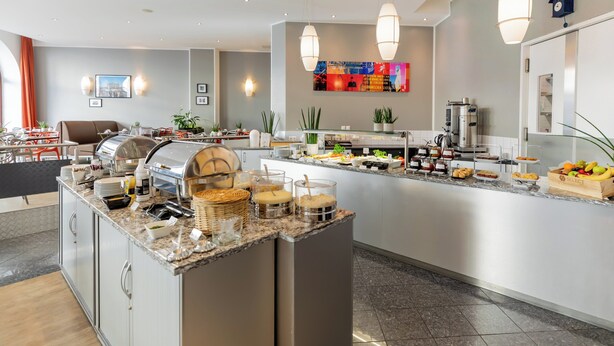 A modern hotel lobby with a central island featuring a buffet, seating area, and a wall-mounted TV. - Garner Hotel Berlin - Checkpoint Charlie by IHG (Berlin)
