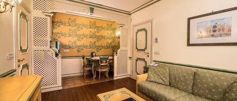 A room with a wooden floor, a sofa, a table with a book, a dining area with chairs, and a decorative wall with a floral pattern.