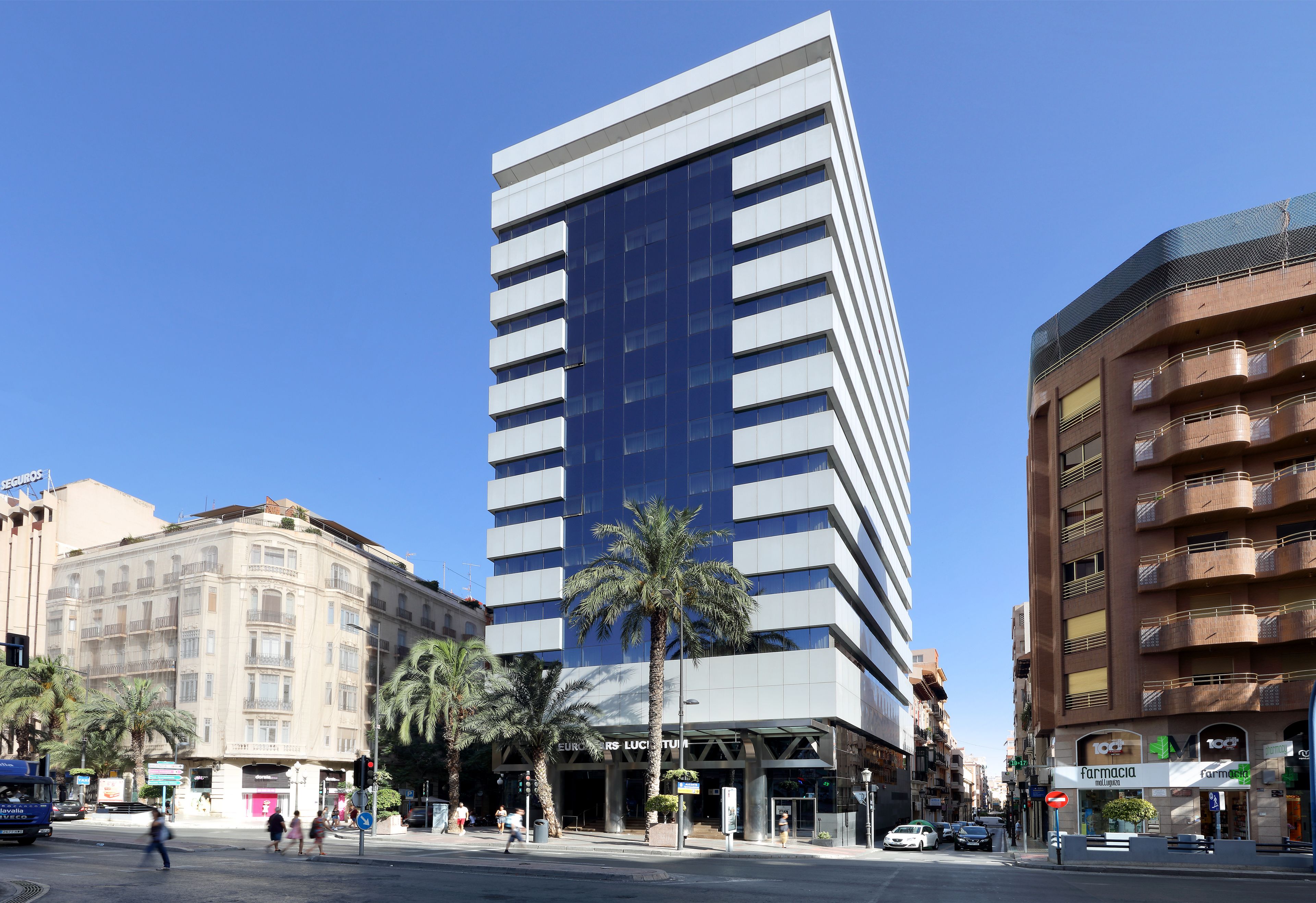 Un edificio moderno con fachada de vidrio, palmeras en primer plano y una calle con coches y peatones.