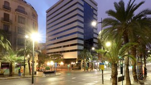 Un edificio moderno con techo plano y varios pisos, rodeado de palmeras y farolas.
