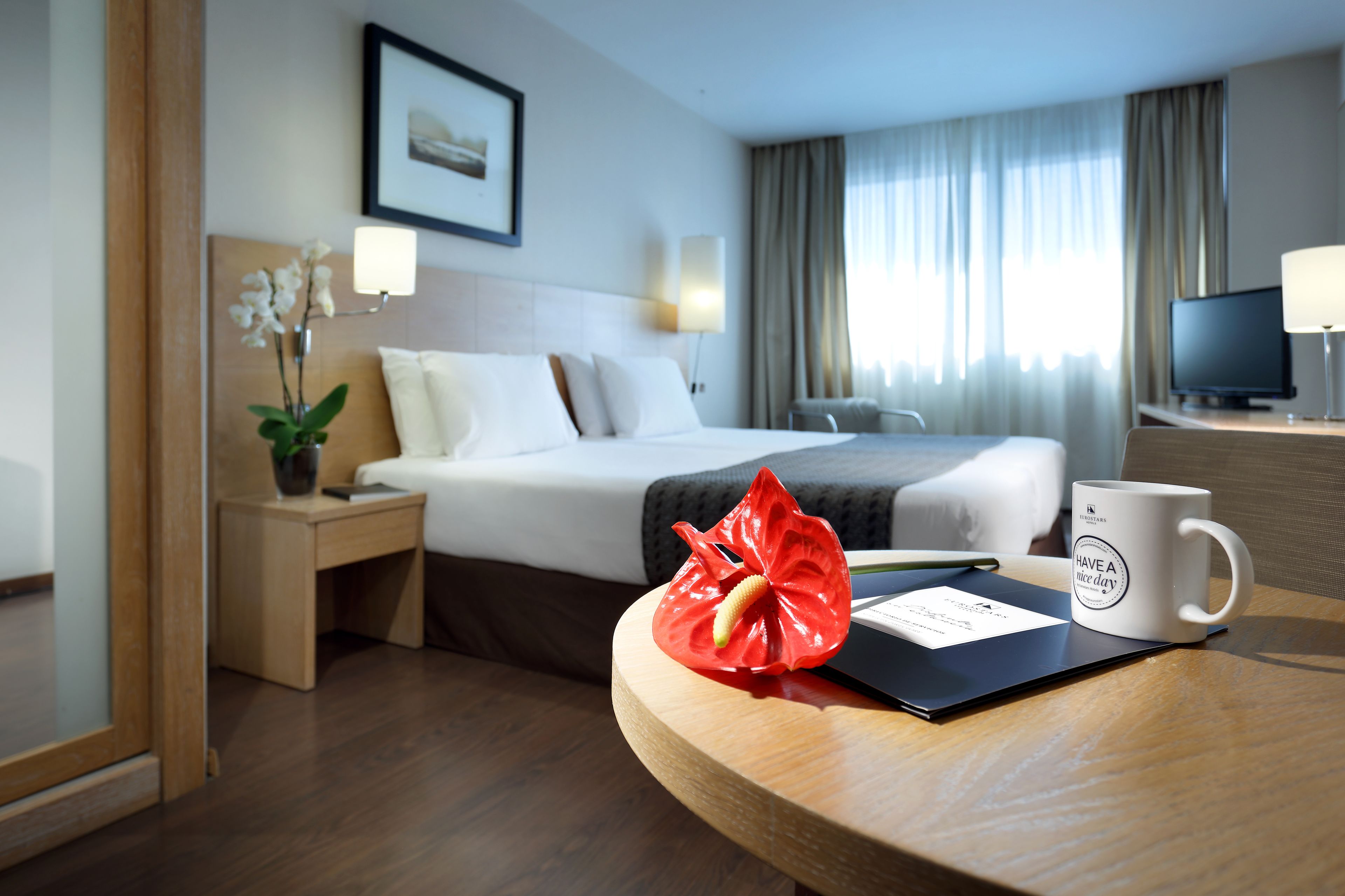 A hotel room with a bed, bedside table, lamp, and a coffee mug on a table.