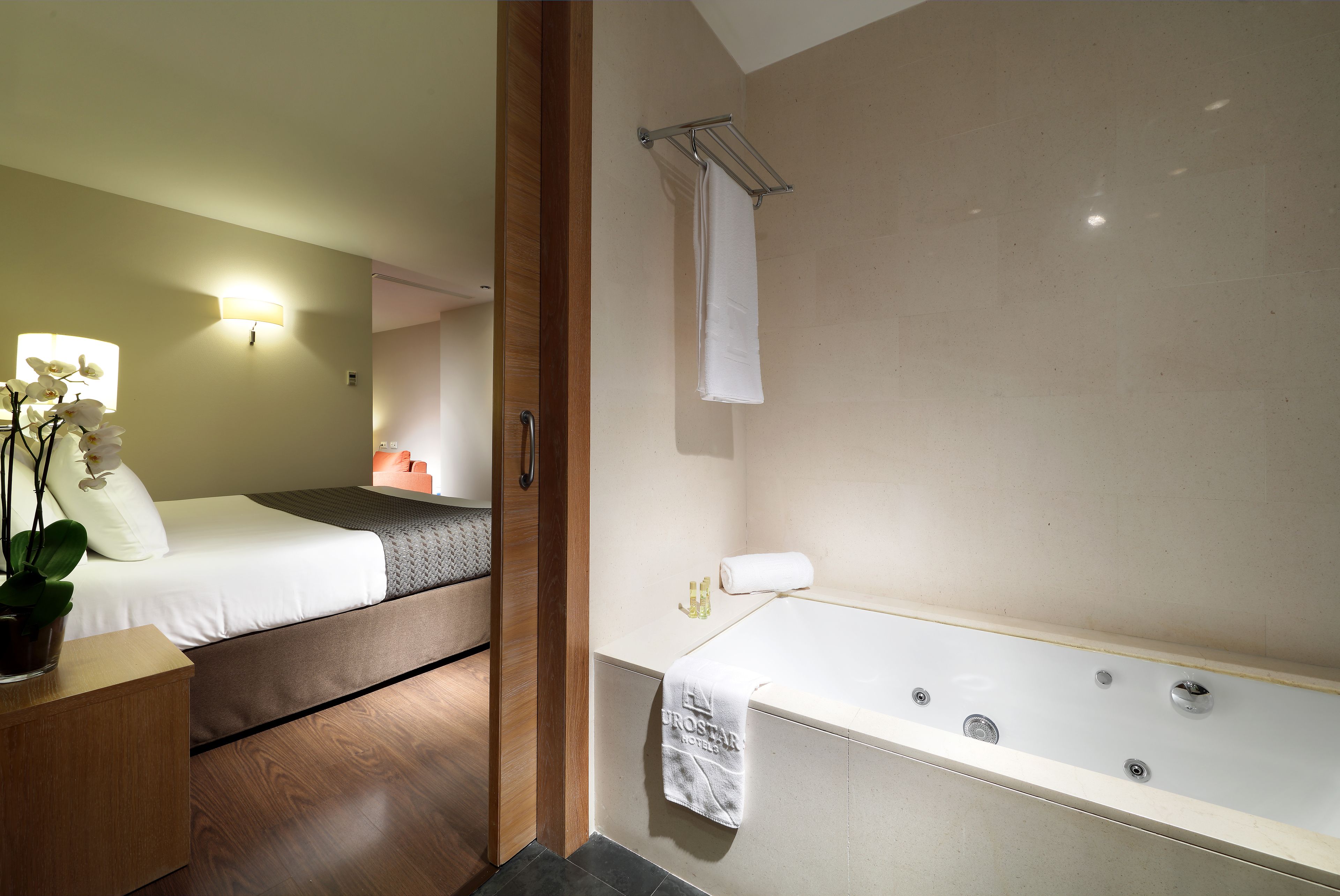 Een moderne hotelkamer met een ruim bad, een bed met een grijze hoofdeinde en een potplant op een houten nachtkastje.