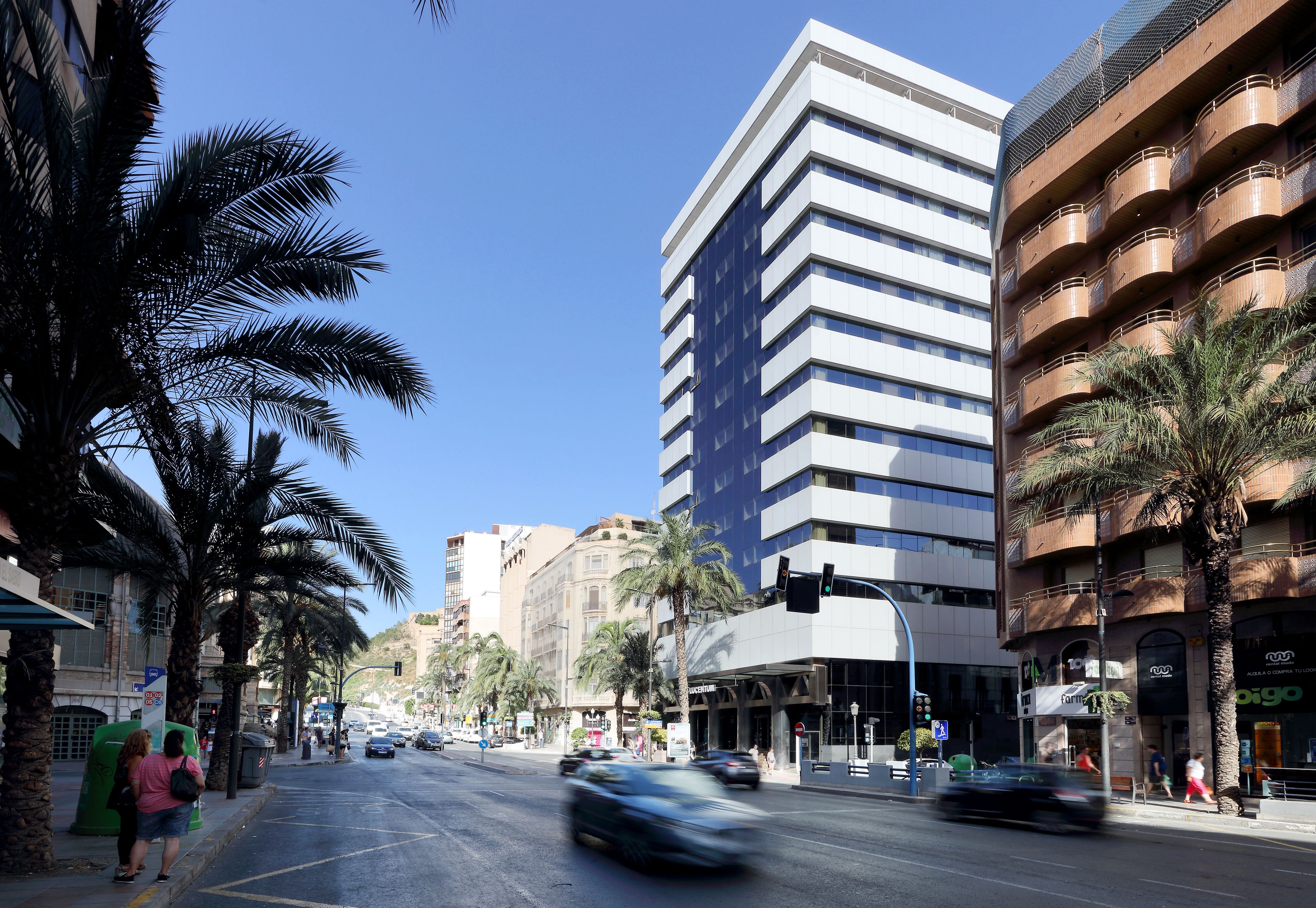 Une rue moderne en milieu urbain, bordée de palmiers, avec un immeuble à plusieurs étages doté de balcons et un carrefour très fréquenté.
