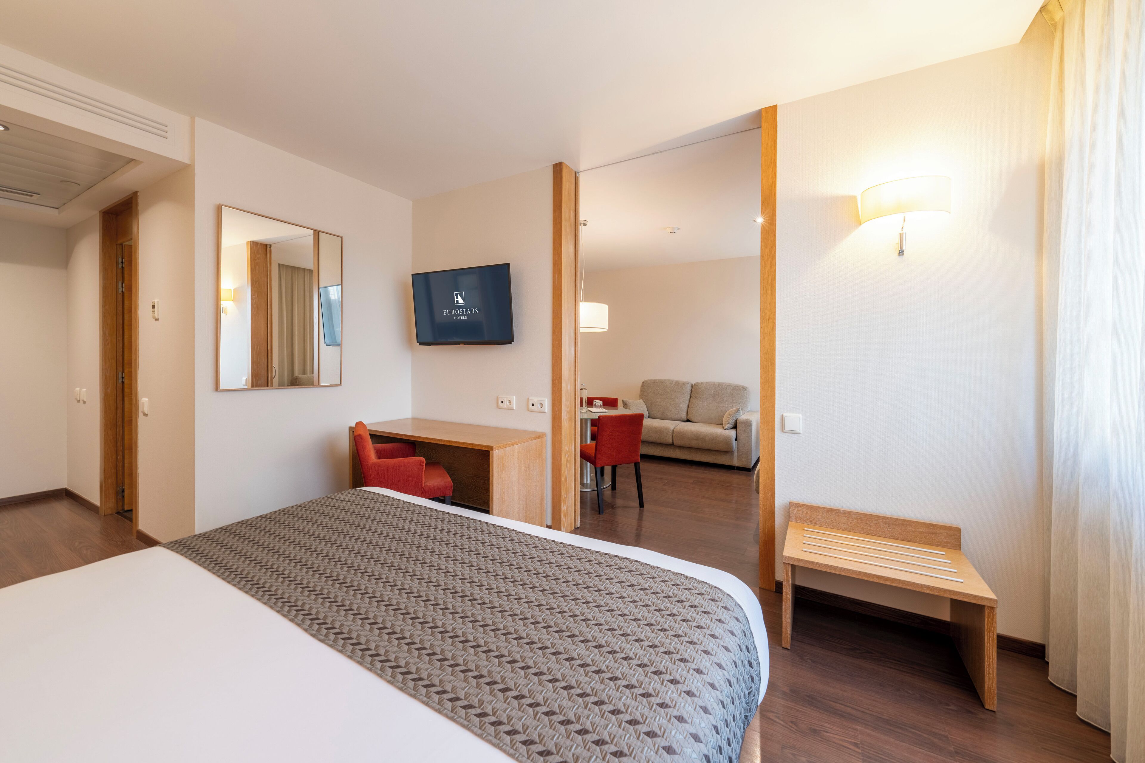 A hotel room with a bed, a desk, a chair, a sofa, a bench, a TV, and a mirror.