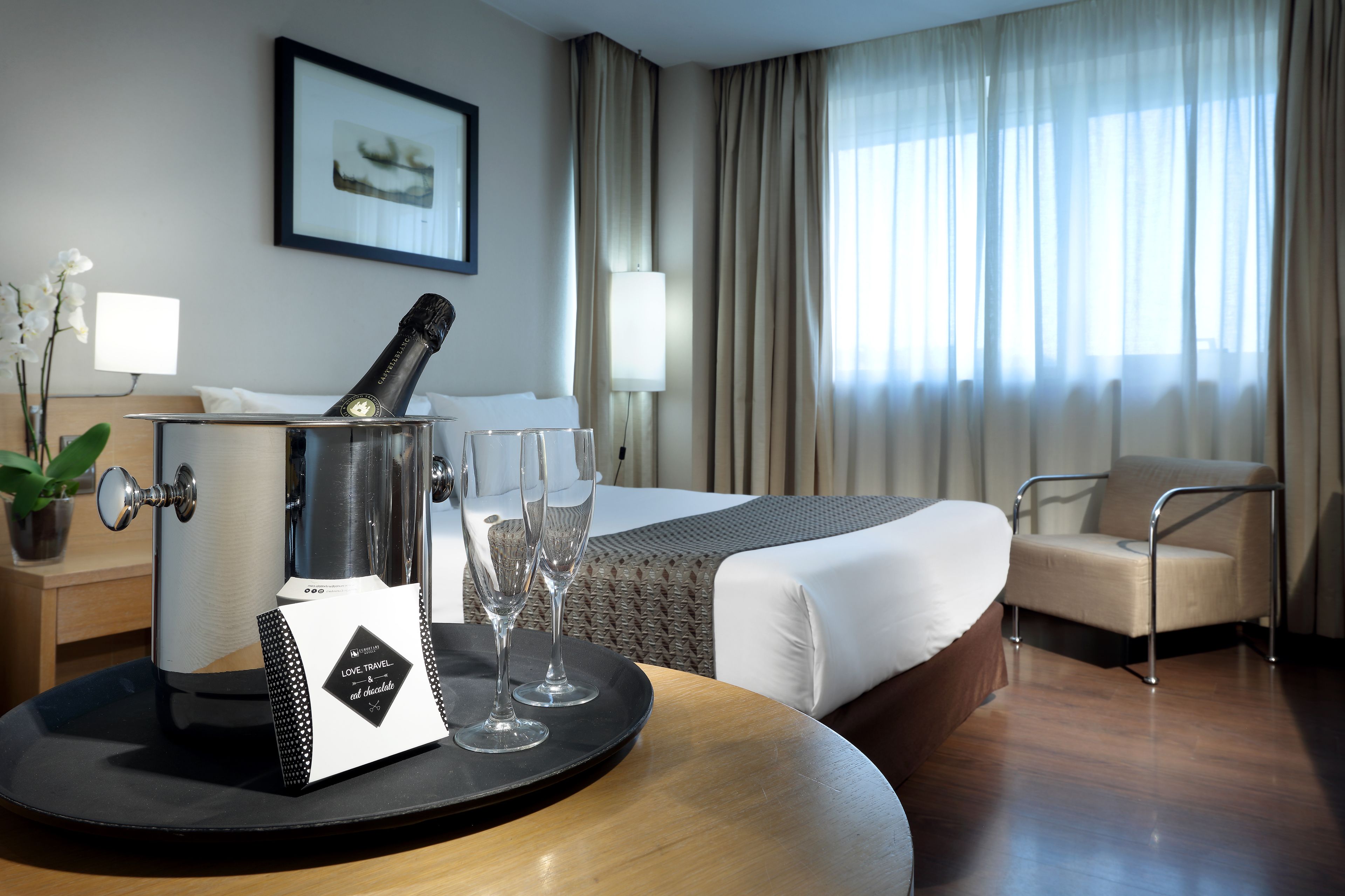 Een hotelkamer met een bed, een stoel, een tafel met een fles en glazen, een lamp en een raam met gordijnen.