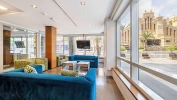 A modern hotel lobby with a large seating area, a flat-screen TV, and large windows offering a view of the cityscape.