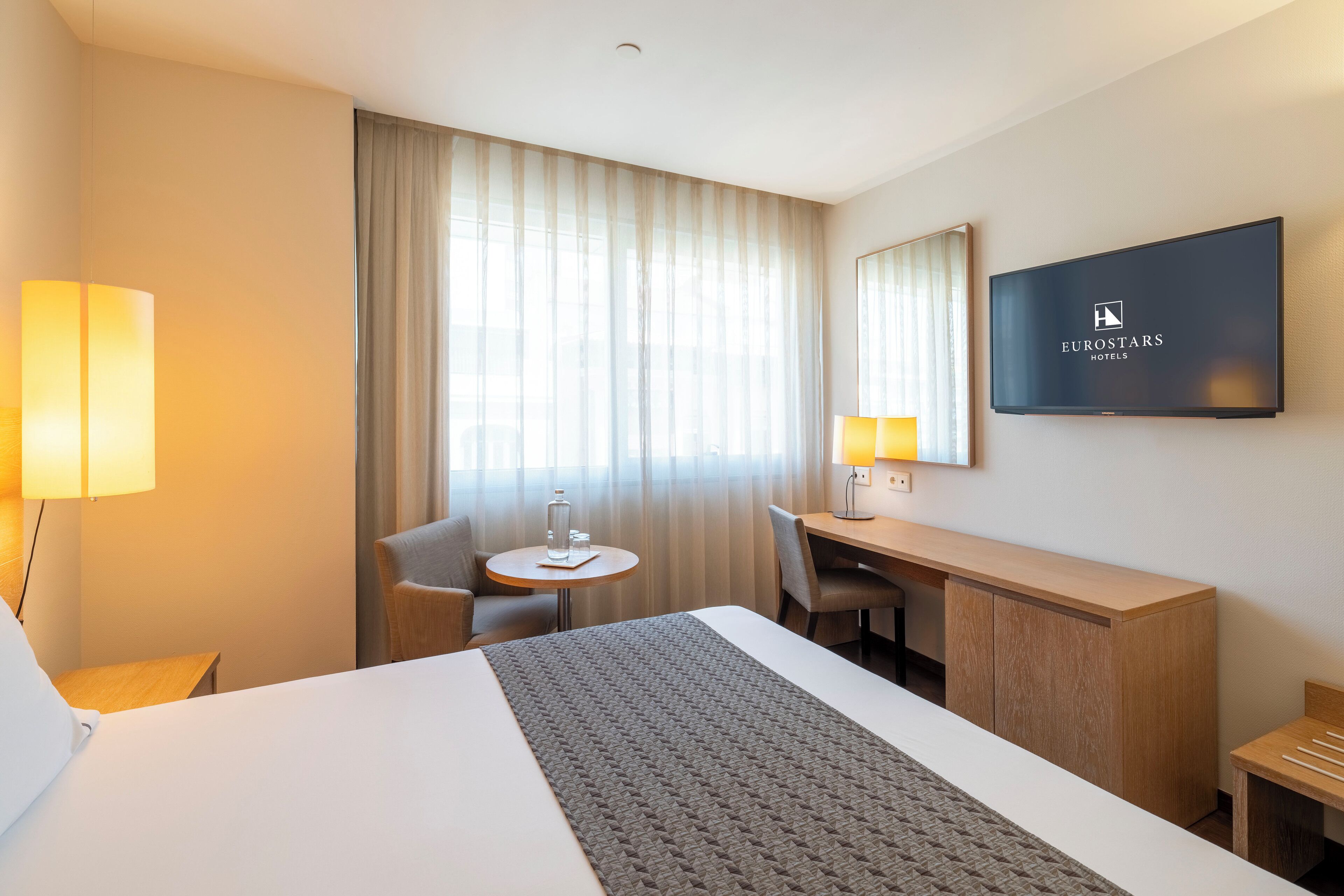 A hotel room with a bed, a desk, a chair, a TV, and a window with curtains.