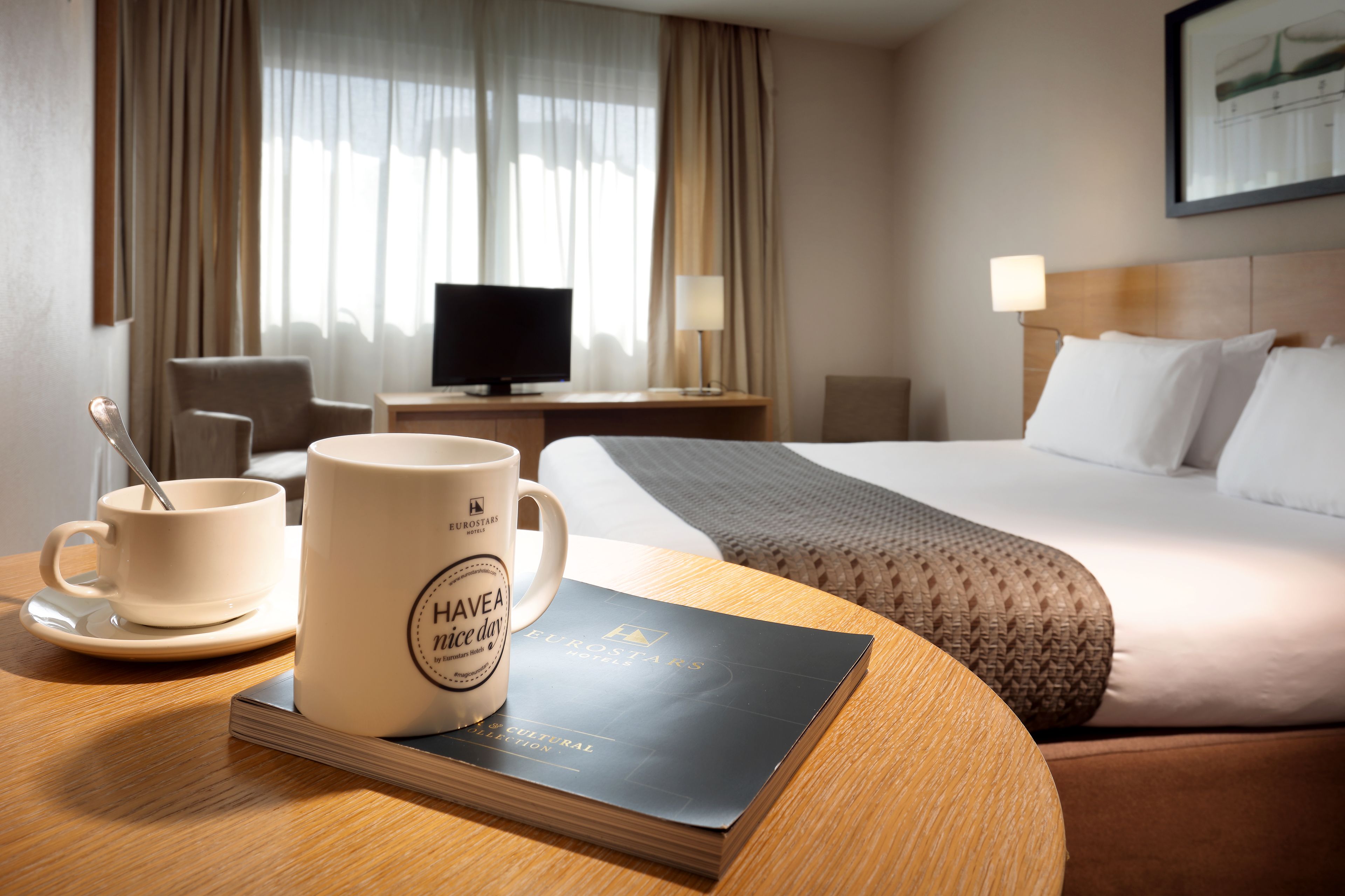 Een hotelkamer met een bed, een nachtkastje met een lamp, een televisie, een koffiekop en een boek op het tafeltje.