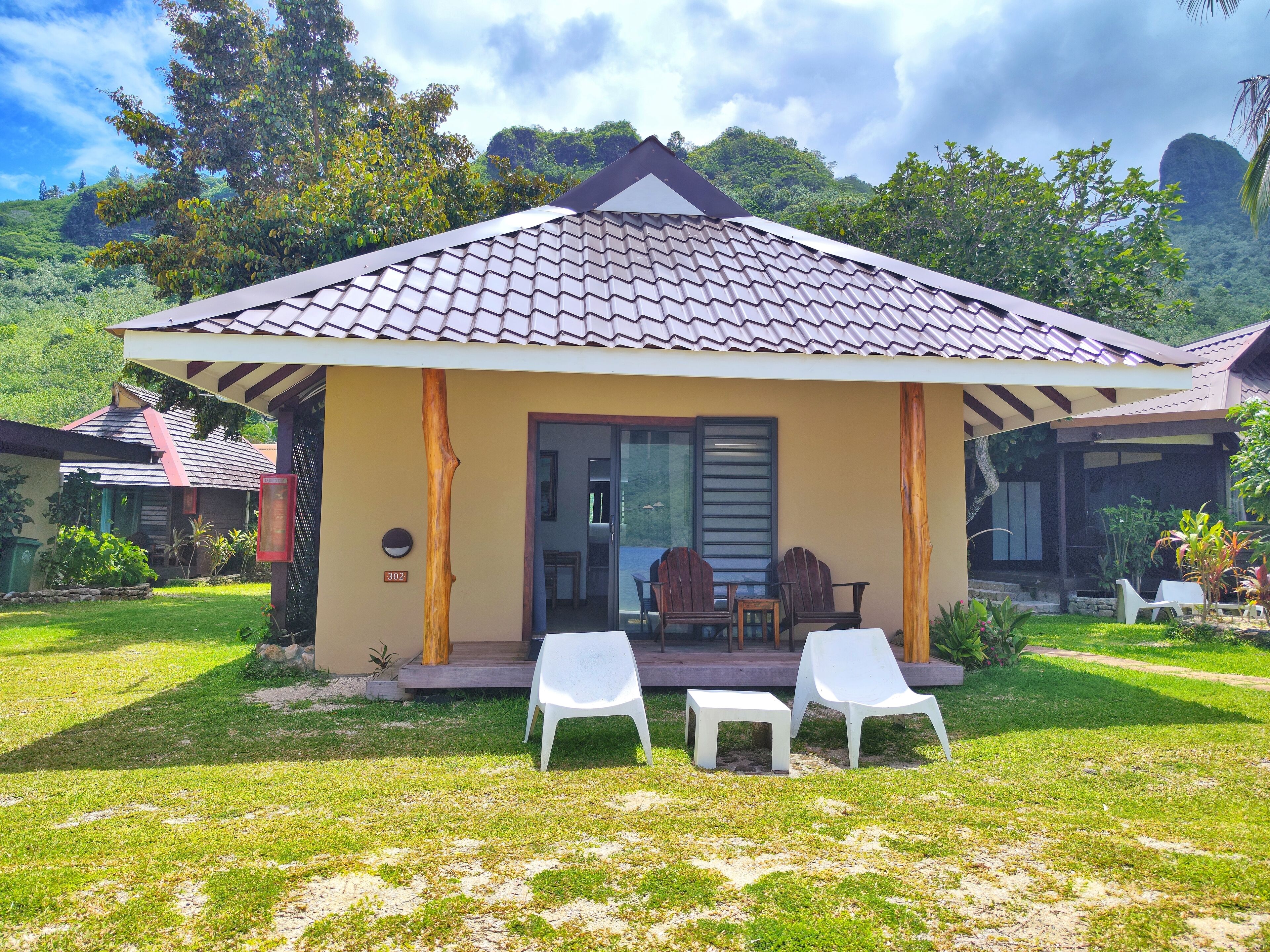 A small, single-story building with a tiled roof and a covered porch. There are two white plastic chairs and a small table on the porch. The building is surrounded by a grassy lawn and has a door leading to an interior space.