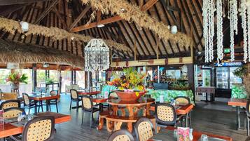 A restaurant with wooden tables and chairs, hanging decorations, and a thatched roof.