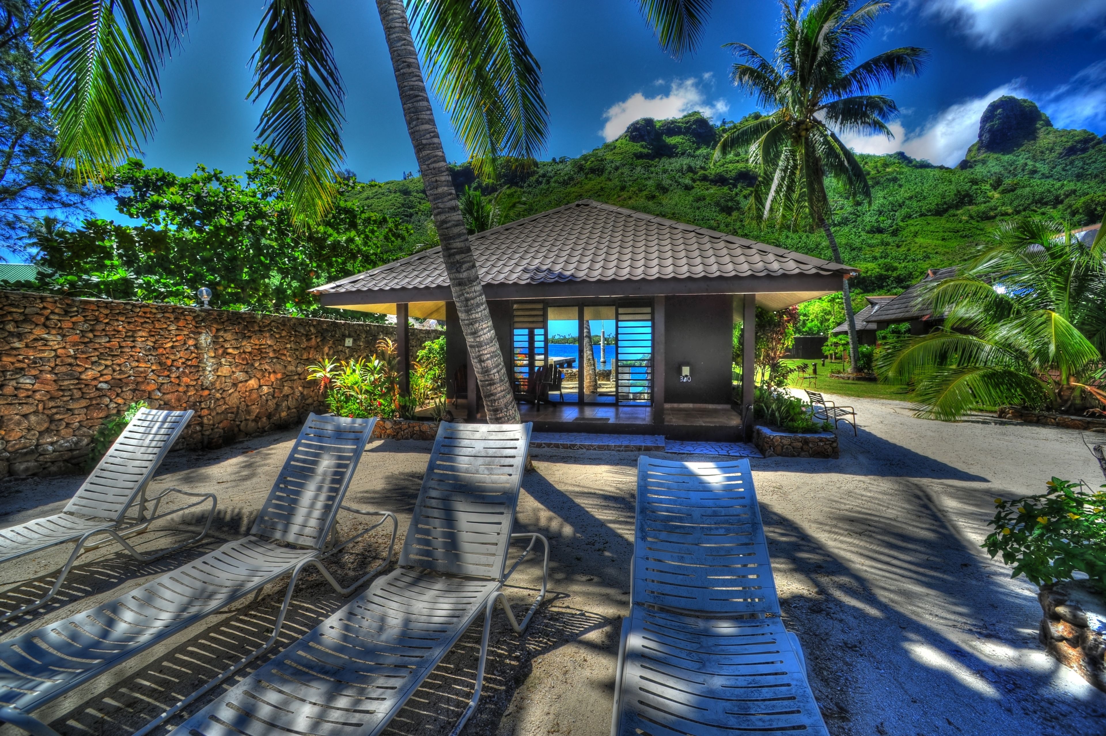 A beachside resort with a stone wall, lounge chairs, a small building with a tiled roof, and lush greenery.