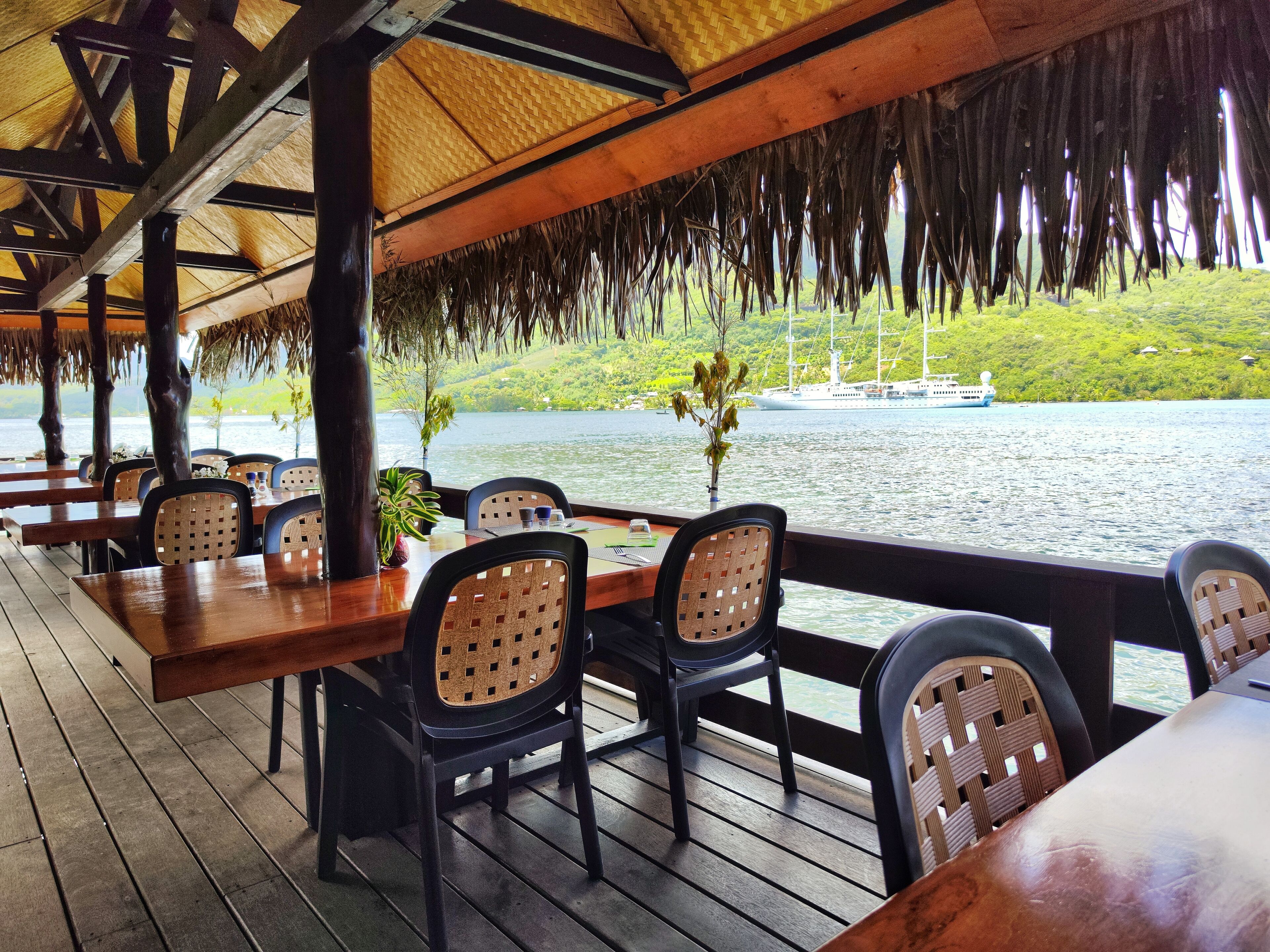 Un espace extérieur aménagé pour les repas, avec des tables et des chaises en bois, donnant sur une étendue d’eau où l’on aperçoit un bateau au loin.