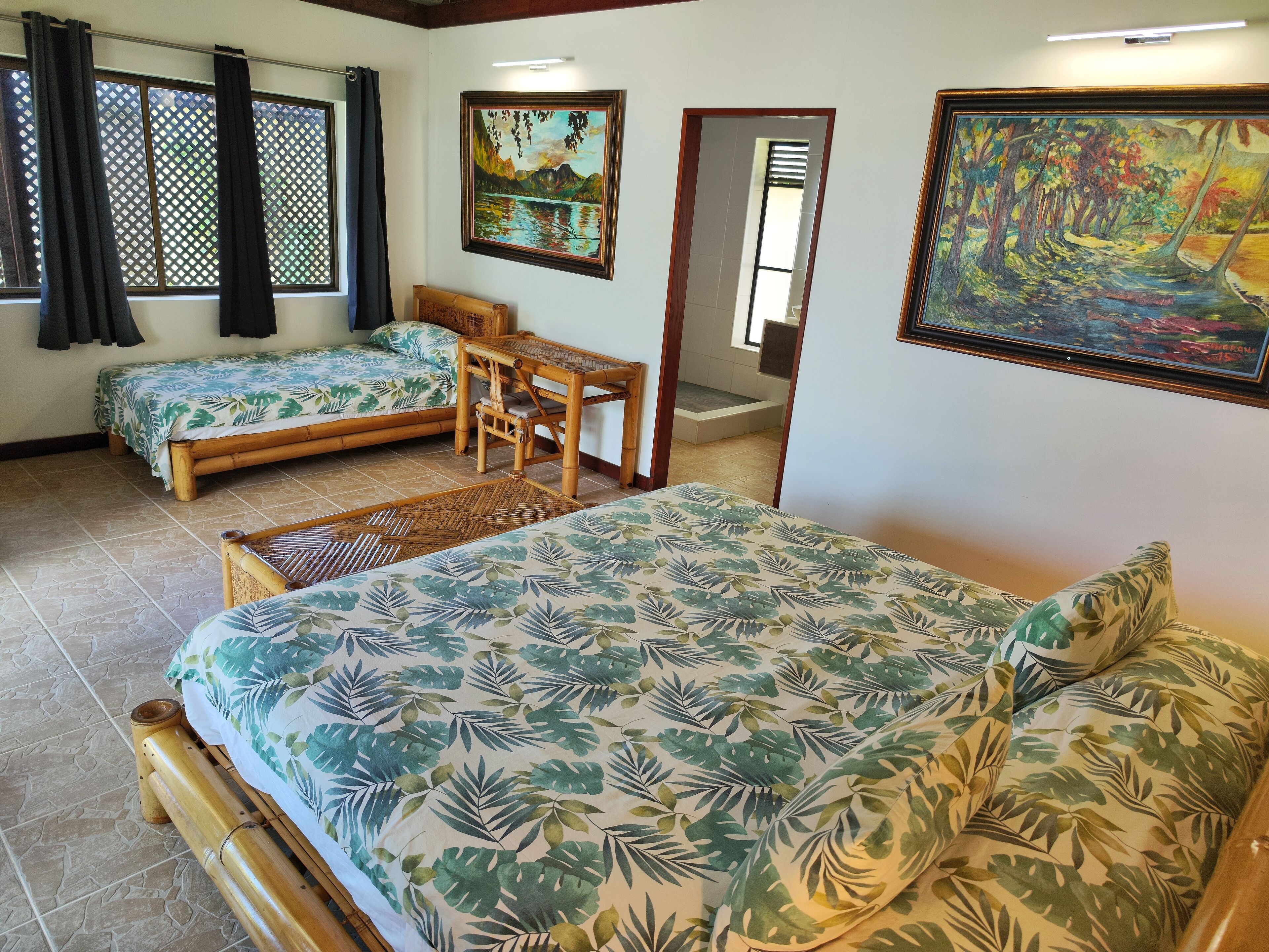 Une chambre avec deux lits simples, une table en bois et une chaise. Il y a deux tableaux accrochés aux murs et une fenêtre avec des rideaux.