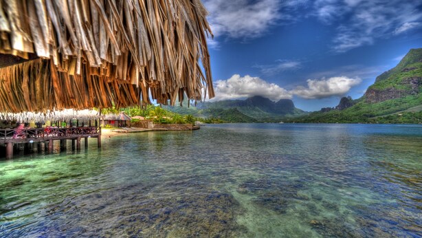 A clear view of a thatched-roof structure extending over a calm body of water, with a mountainous landscape in the background. - Hotel Kaveka (Moorea-Maiao)