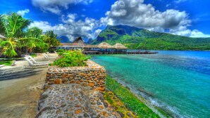 A beach resort with thatched-roof overwater bungalows, a stone wall, and a pier extending into turquoise waters. - Hotel Kaveka (Moorea-Maiao)