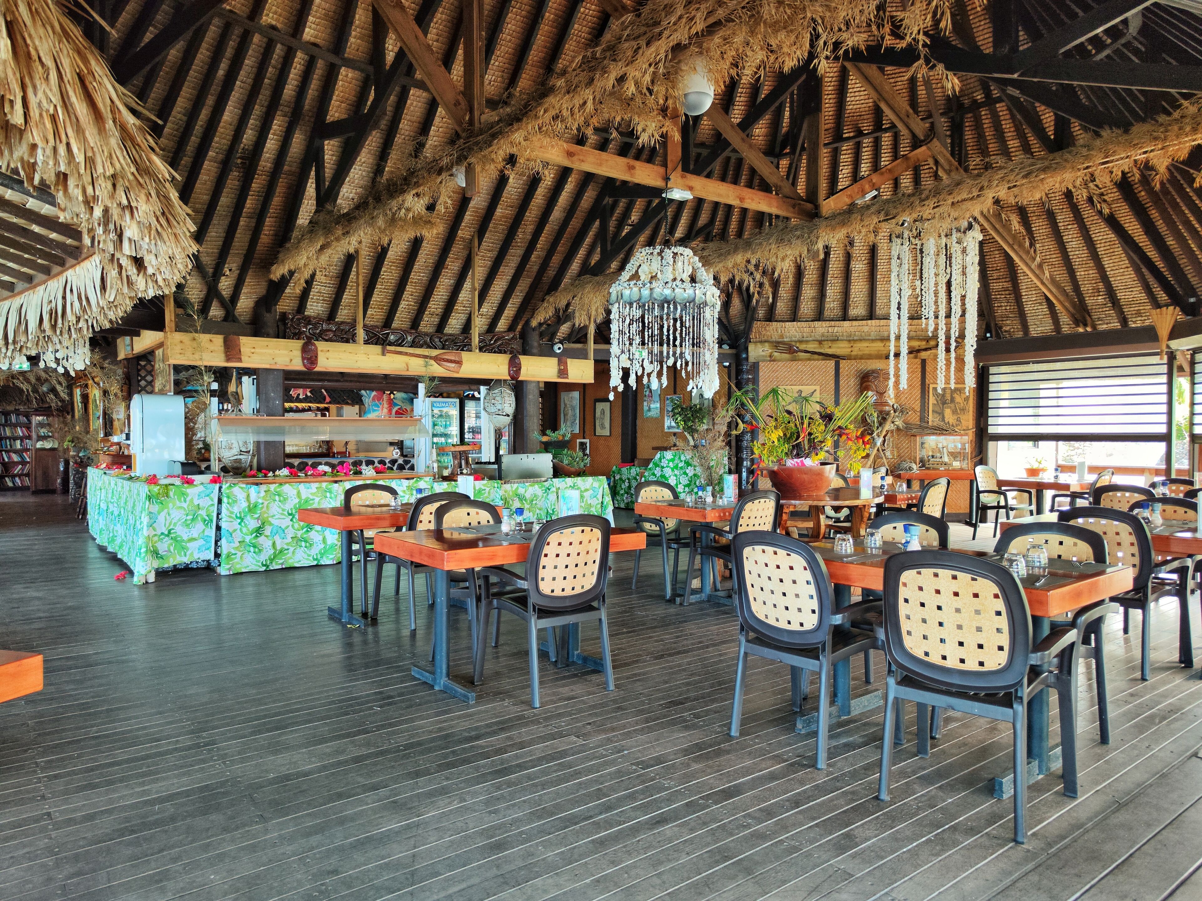 A restaurant with a thatched roof, wooden beams, and a bar area with a green floral tablecloth.
