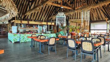 A restaurant with a thatched roof, wooden beams, and a bar area with a green floral tablecloth.