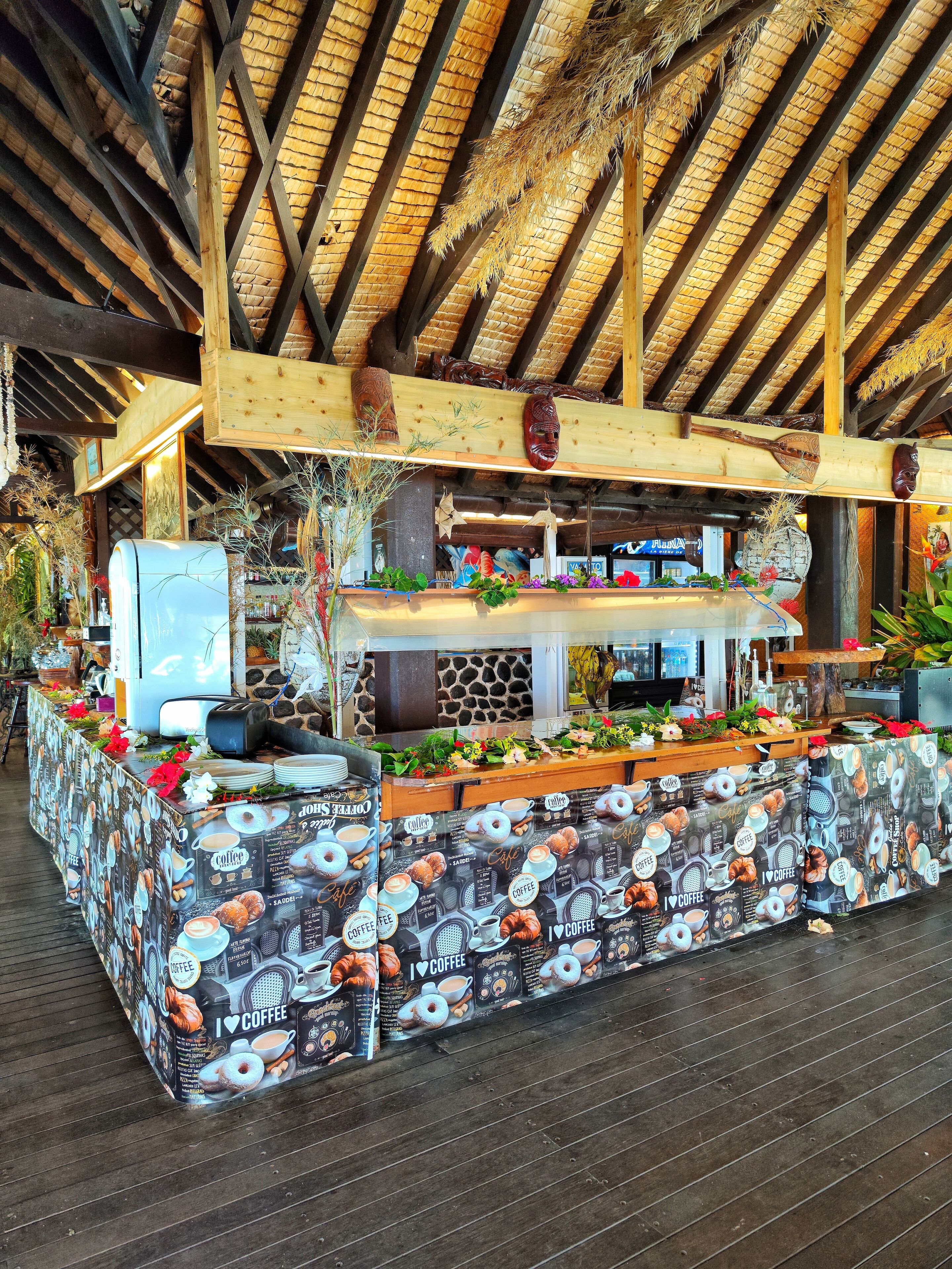 A rustic coffee shop with a thatched roof, wooden beams, and a counter decorated with coffee-themed items and plants.