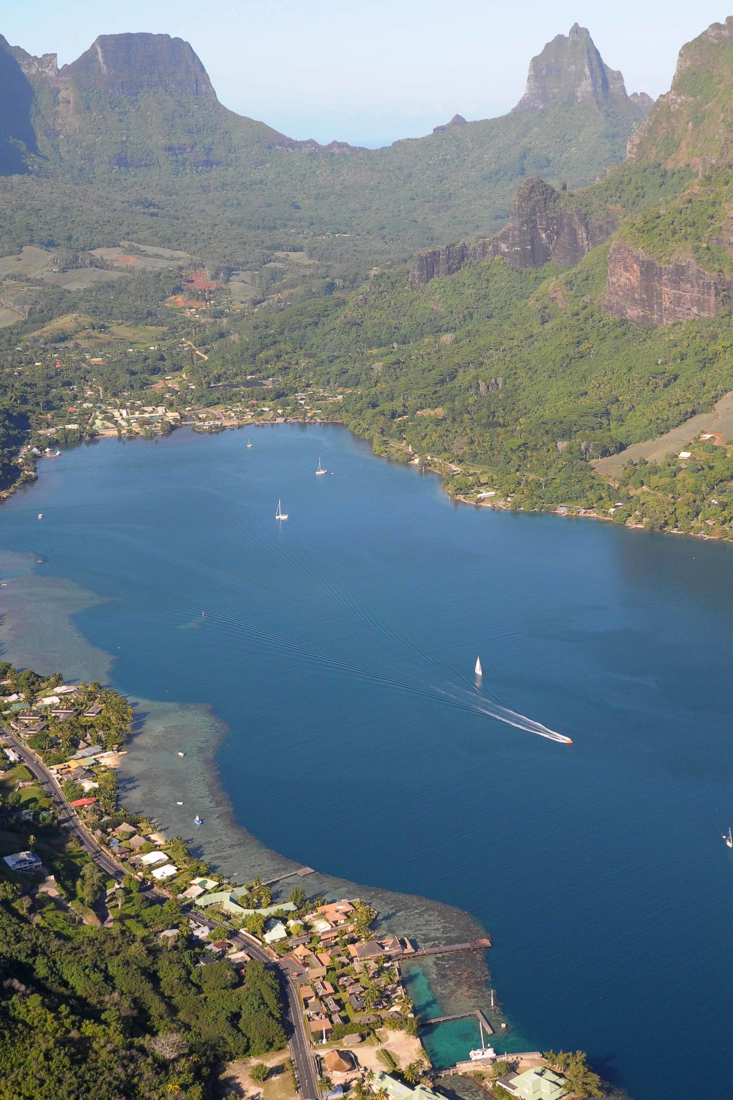An aerial view of a coastal town with a large body of water, several boats, and surrounding green hills.