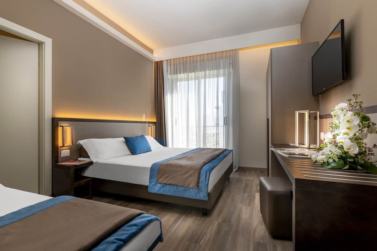 A hotel room with two beds, a desk with a flat-screen TV, a nightstand with a lamp, and a large window with sheer curtains.