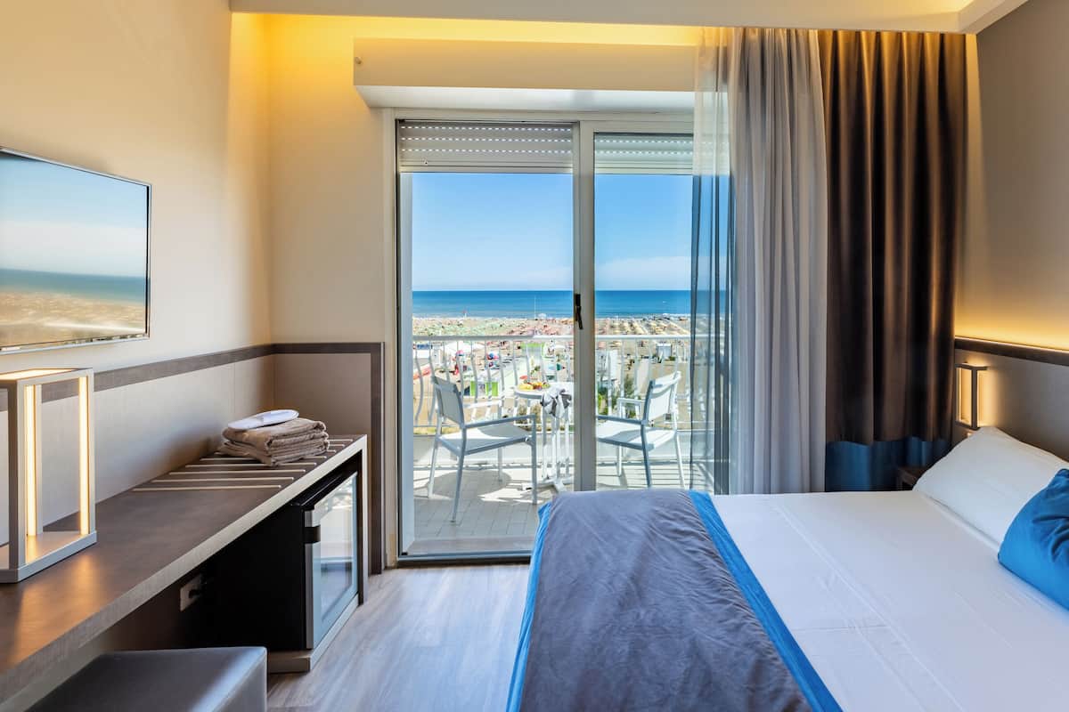 A hotel room with a bed, a desk, a chair, a TV, and a balcony with a view of the beach.