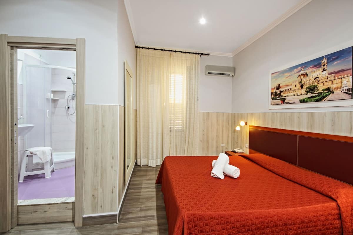 Habitación de hotel con cama, ducha, un taburete pequeño, un cuadro y un equipo de aire acondicionado.