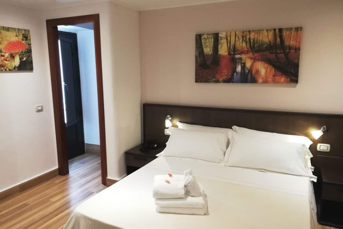 Habitación de hotel con una cama grande, ropa de cama blanca, una mesita de noche, un cuadro con una escena de bosque y piso de madera.