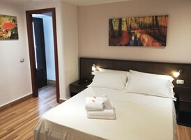 Habitación de hotel con una cama grande, ropa de cama blanca, una mesita de noche, un cuadro con una escena de bosque y piso de madera.