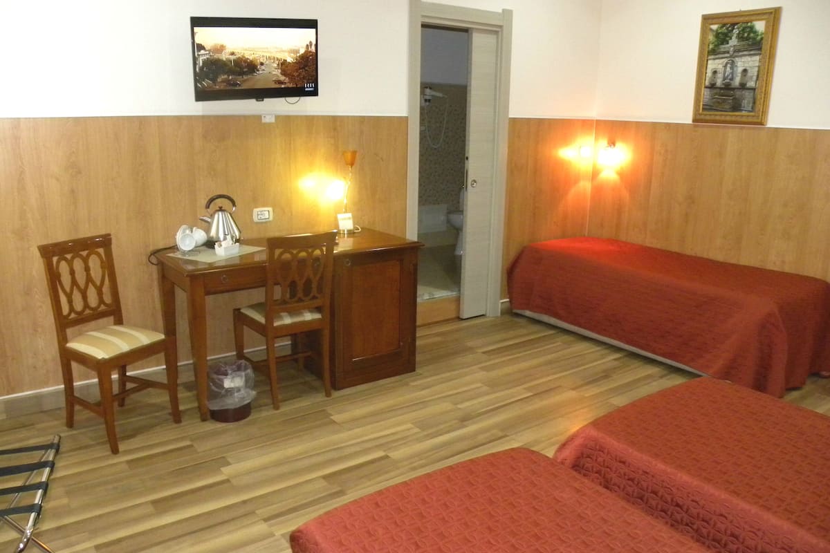 Una habitación con un escritorio de madera, dos sillas, una cama con una manta roja, un televisor en la pared y una puerta que da a otra habitación.