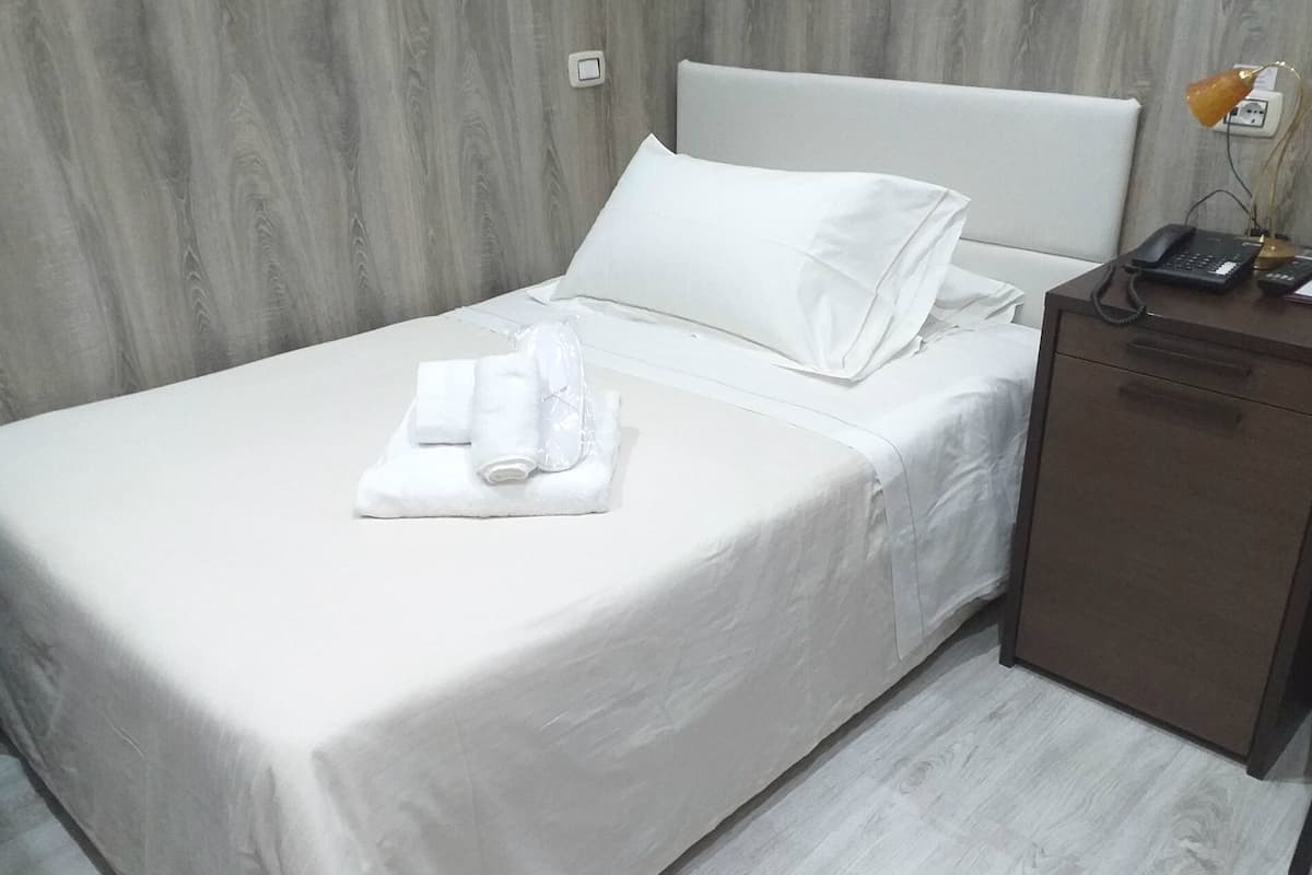 Una cama bien tendida con sábanas blancas, una mesita de noche con una lámpara y un pequeño armario de madera.