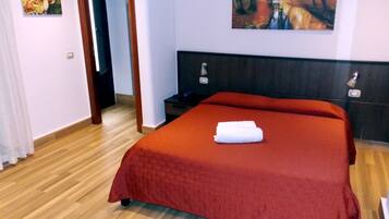 A hotel room with a large bed, wooden flooring, a nightstand, a TV, and two paintings on the walls.