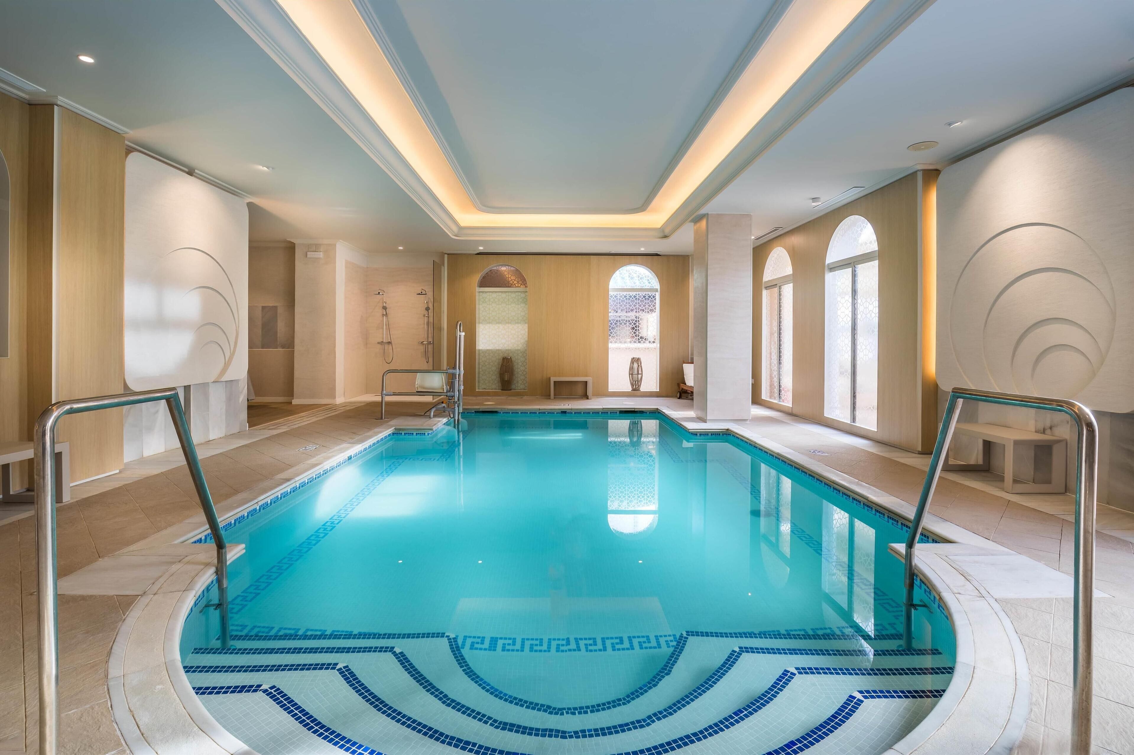 A large indoor swimming pool with clear blue water, surrounded by beige walls and a tiled floor.