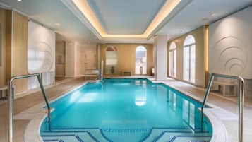 Uma ampla piscina interior com água azul cristalina, rodeada por paredes bege e chão de azulejos.