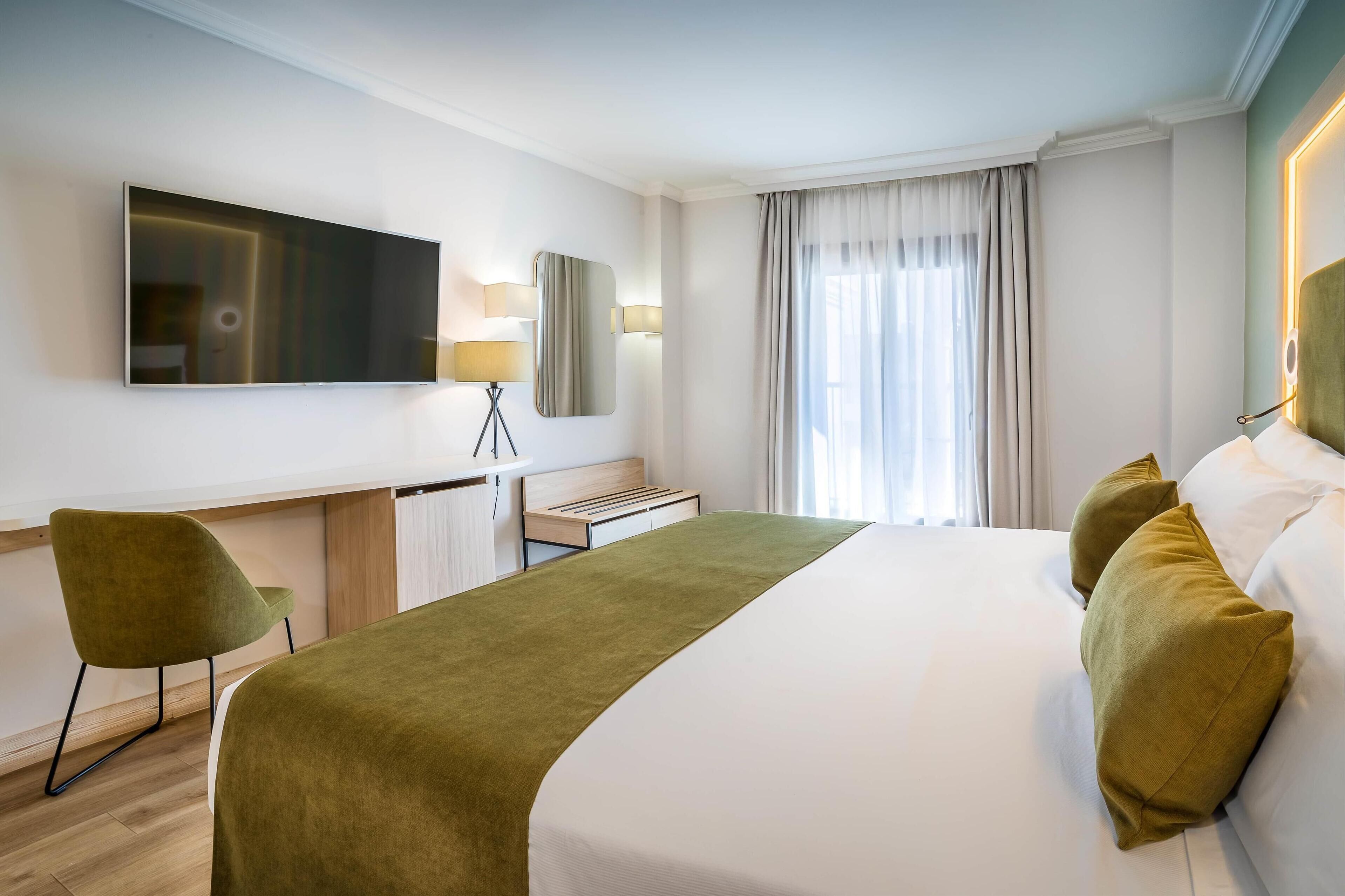 A hotel room with a bed, a desk, a chair, a TV, and a window with curtains.