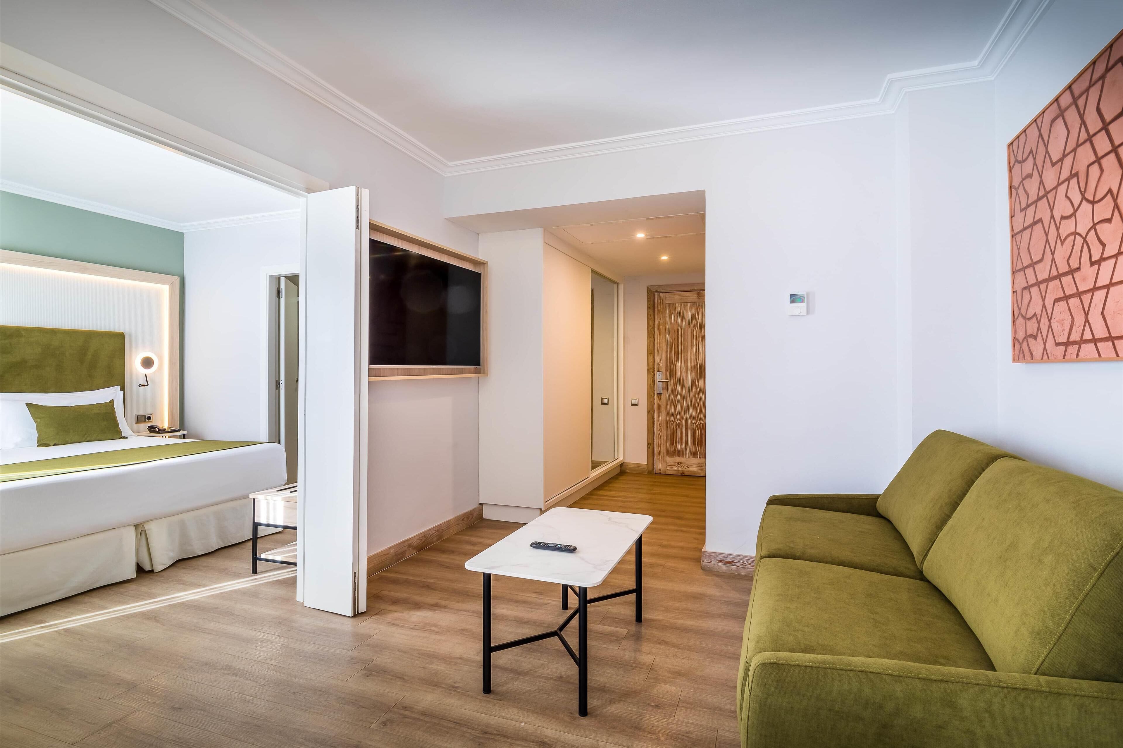A modern hotel room with a bed, a small table, a green sofa, and a TV.