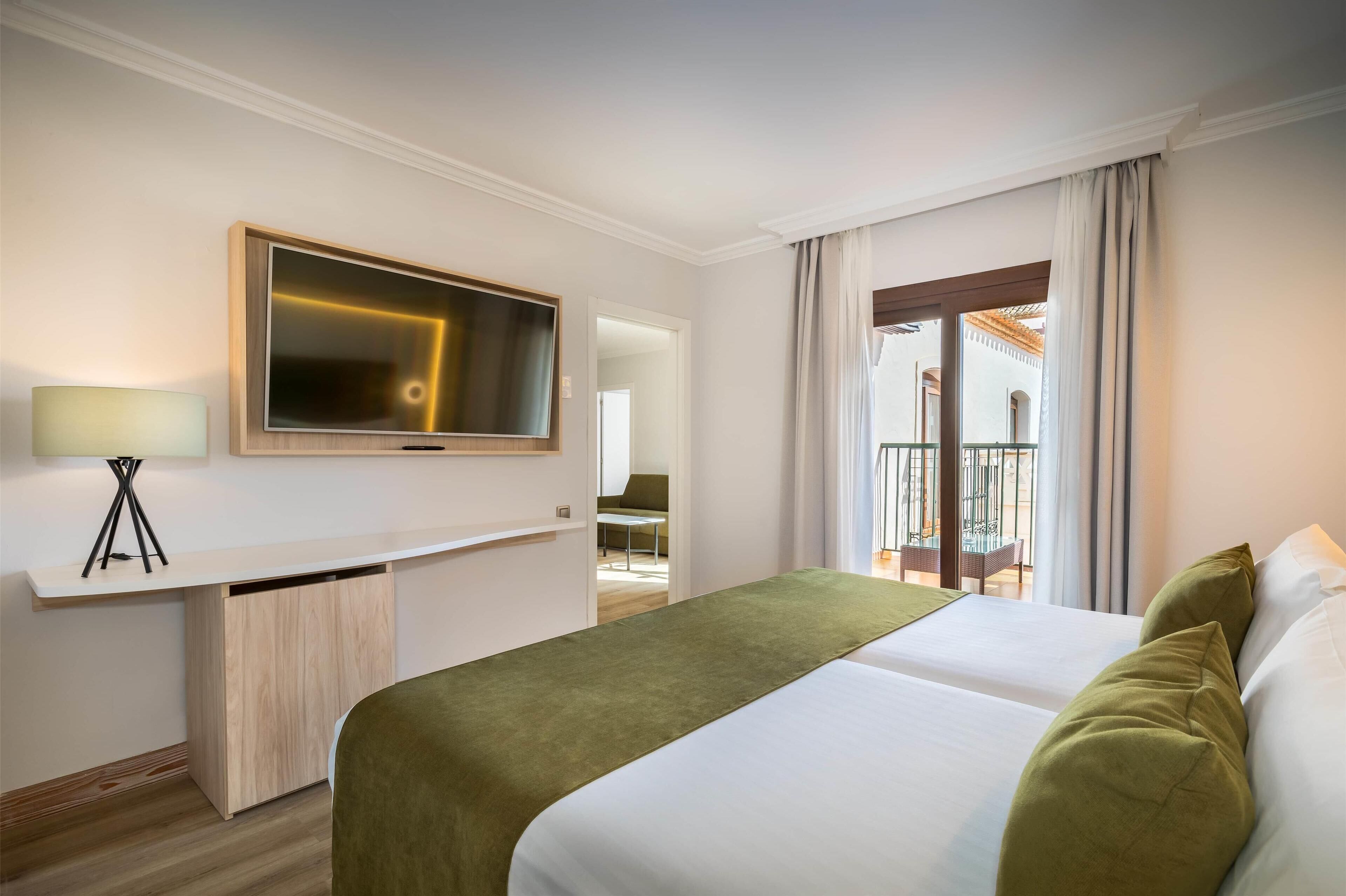 Ein Hotelzimmer mit Bett, Schreibtisch mit Lampe, Fernseher und Balkon mit Stuhl.