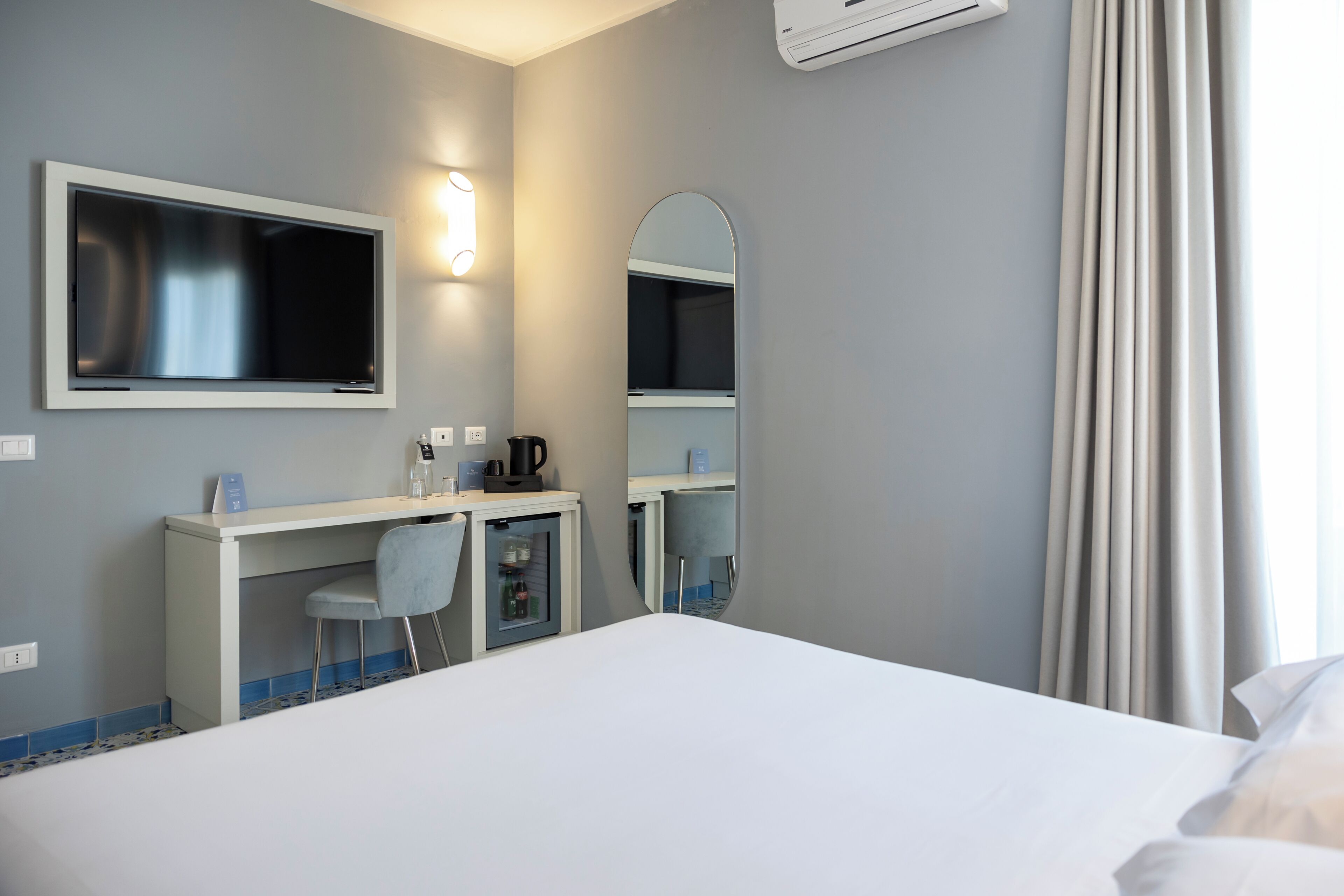 A hotel room with a bed, a desk with a chair, a mirror, and a TV.