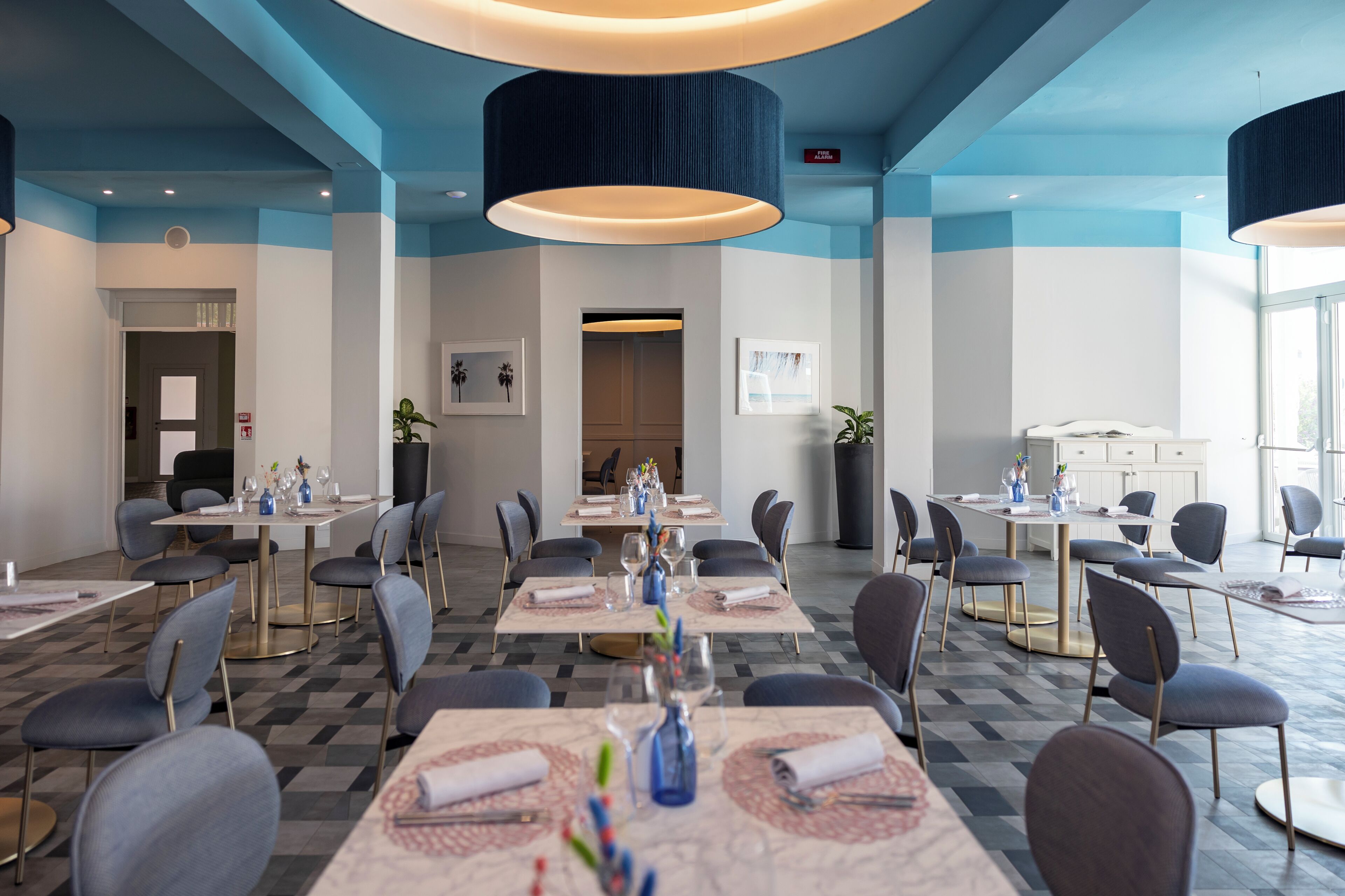 Espaço de refeições de hotel com várias mesas, cadeiras e luminárias pendentes.