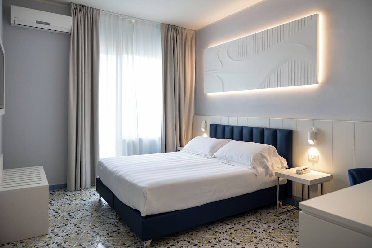 Ein modernes Hotelzimmer mit einem großen Bett, einem Nachttisch mit Lampe, einer Wandleuchte und einem Fenster mit Vorhängen.