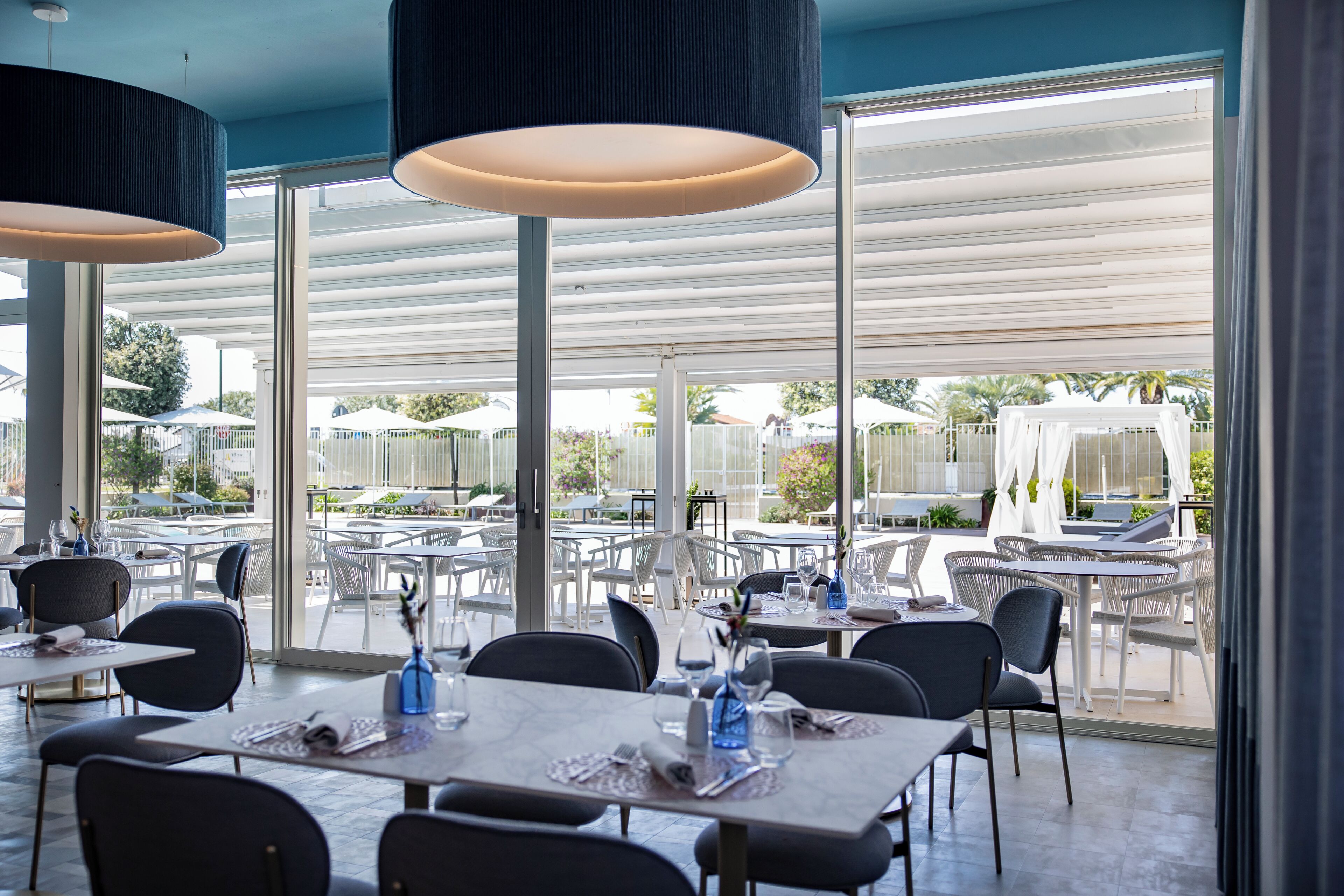 Un comedor interior con mesas y sillas, ventanas amplias y lámparas colgantes.