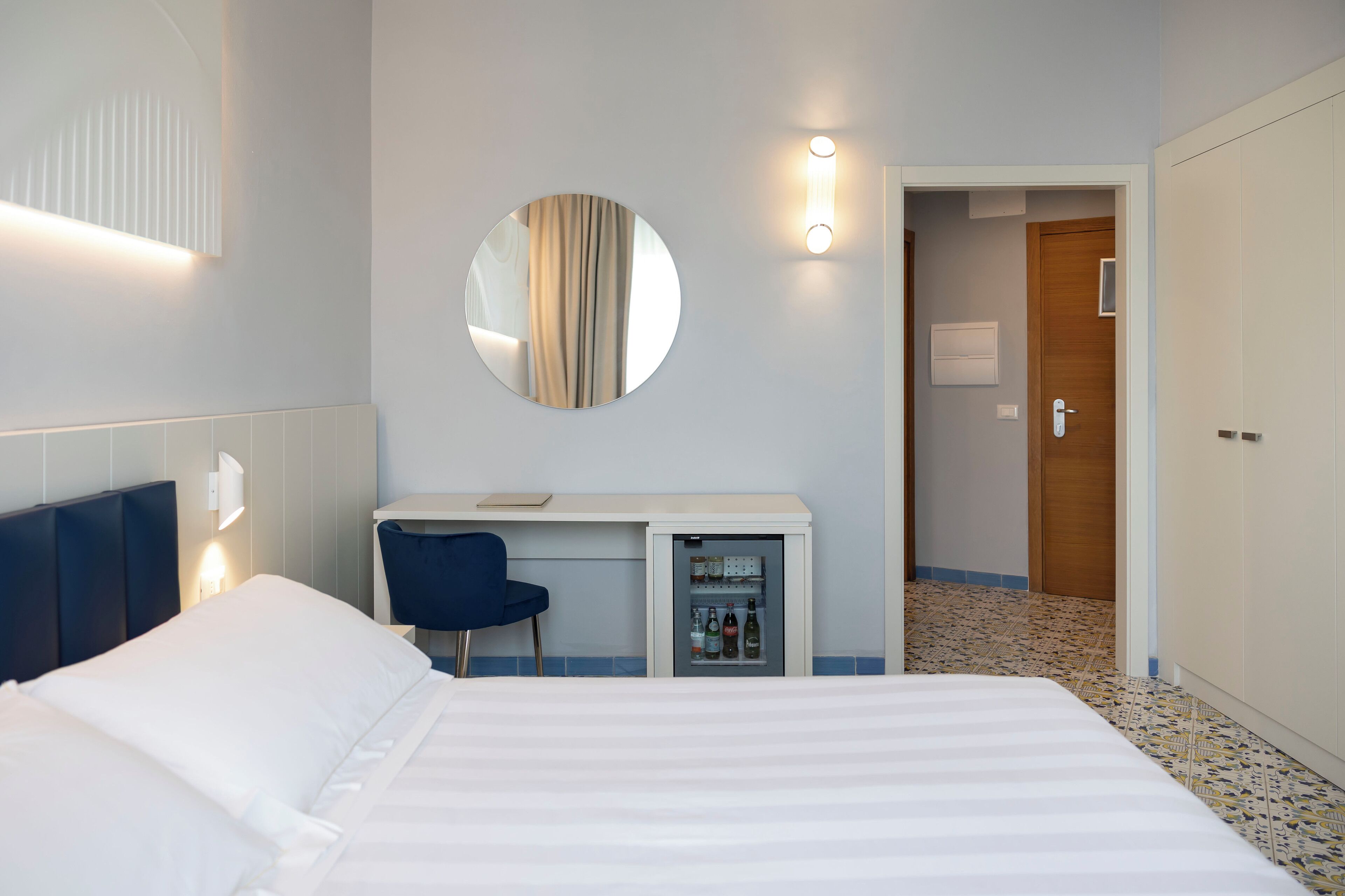 Ein Hotelzimmer mit einem Bett, einem Schreibtisch mit Stuhl, einer Minibar, einem runden Spiegel und einer Tür zu einem weiteren Raum.