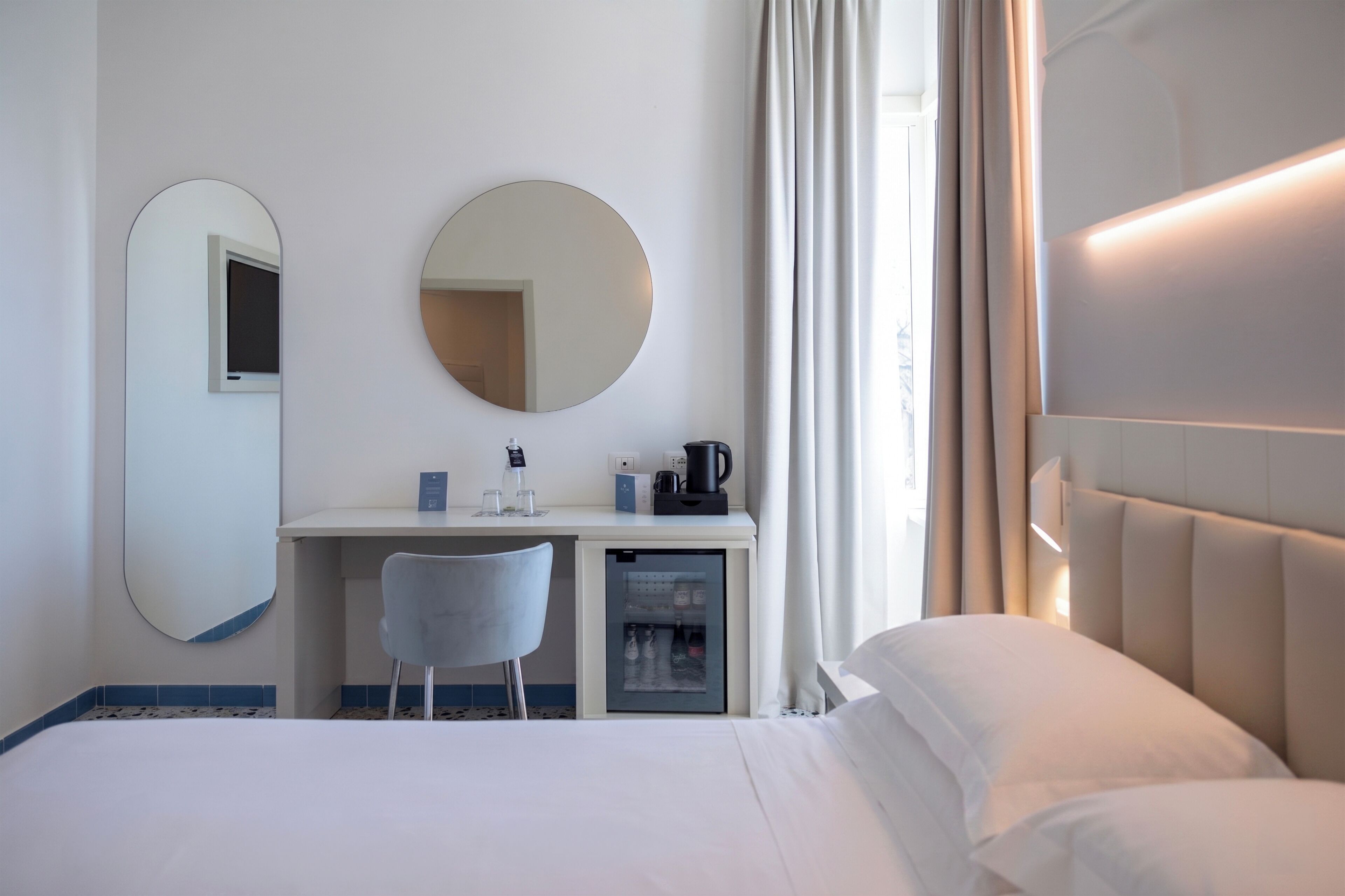A hotel room with a bed, a desk with a mirror, a chair, a small refrigerator, and a coffee maker.