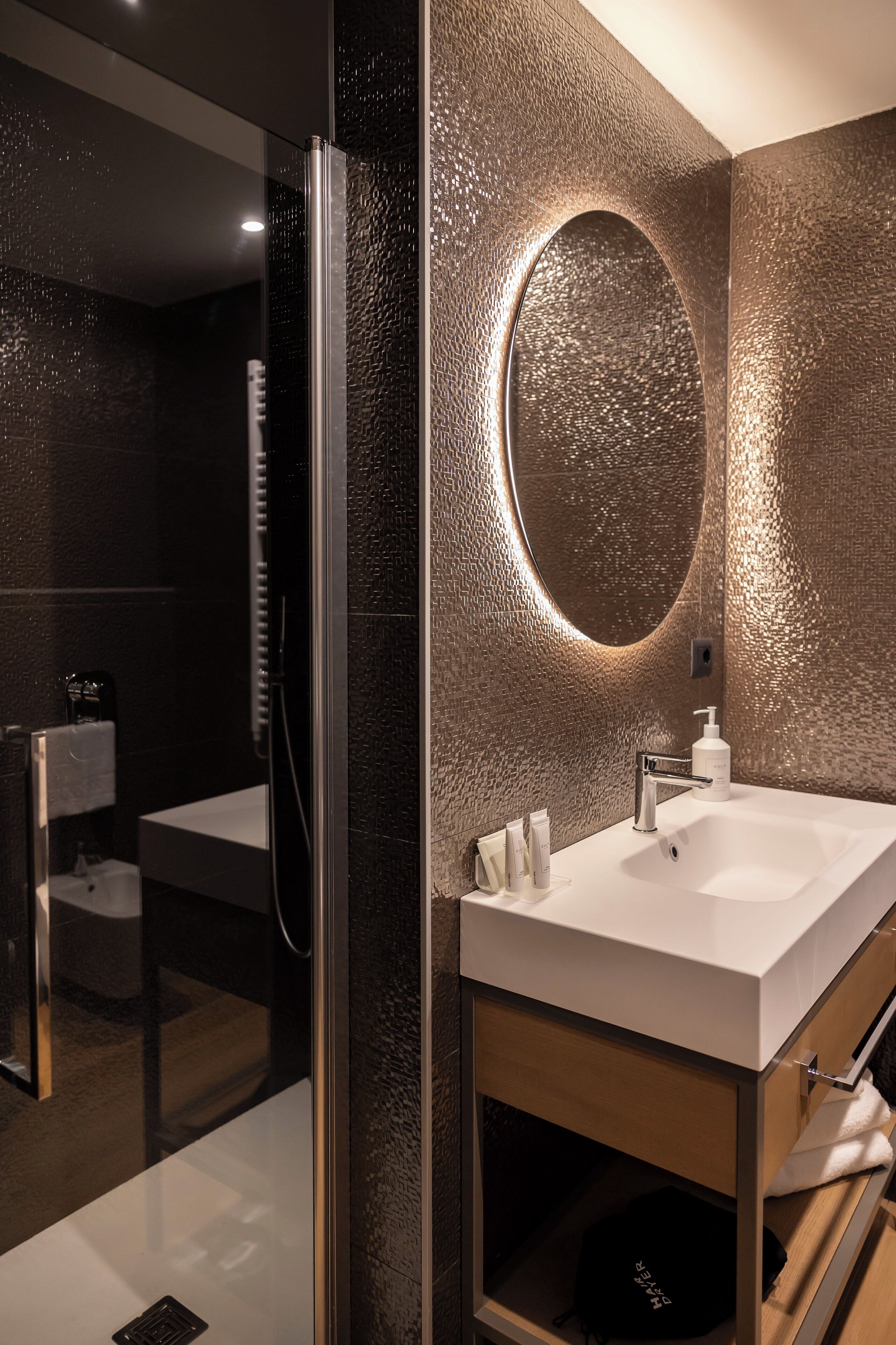 A modern bathroom with a glass-enclosed shower, a round mirror, a white sink, and a wooden cabinet.