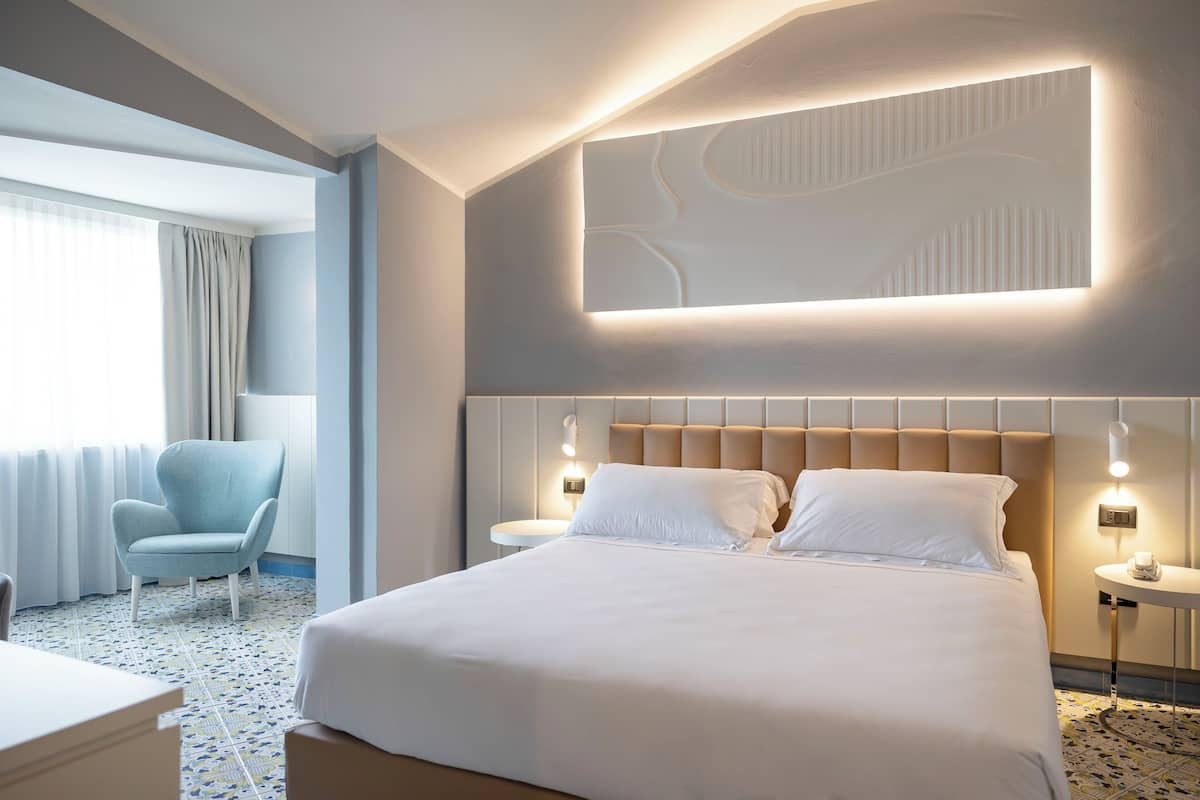 A hotel room with a large bed, a chair, a bedside table, and a wall-mounted light fixture.