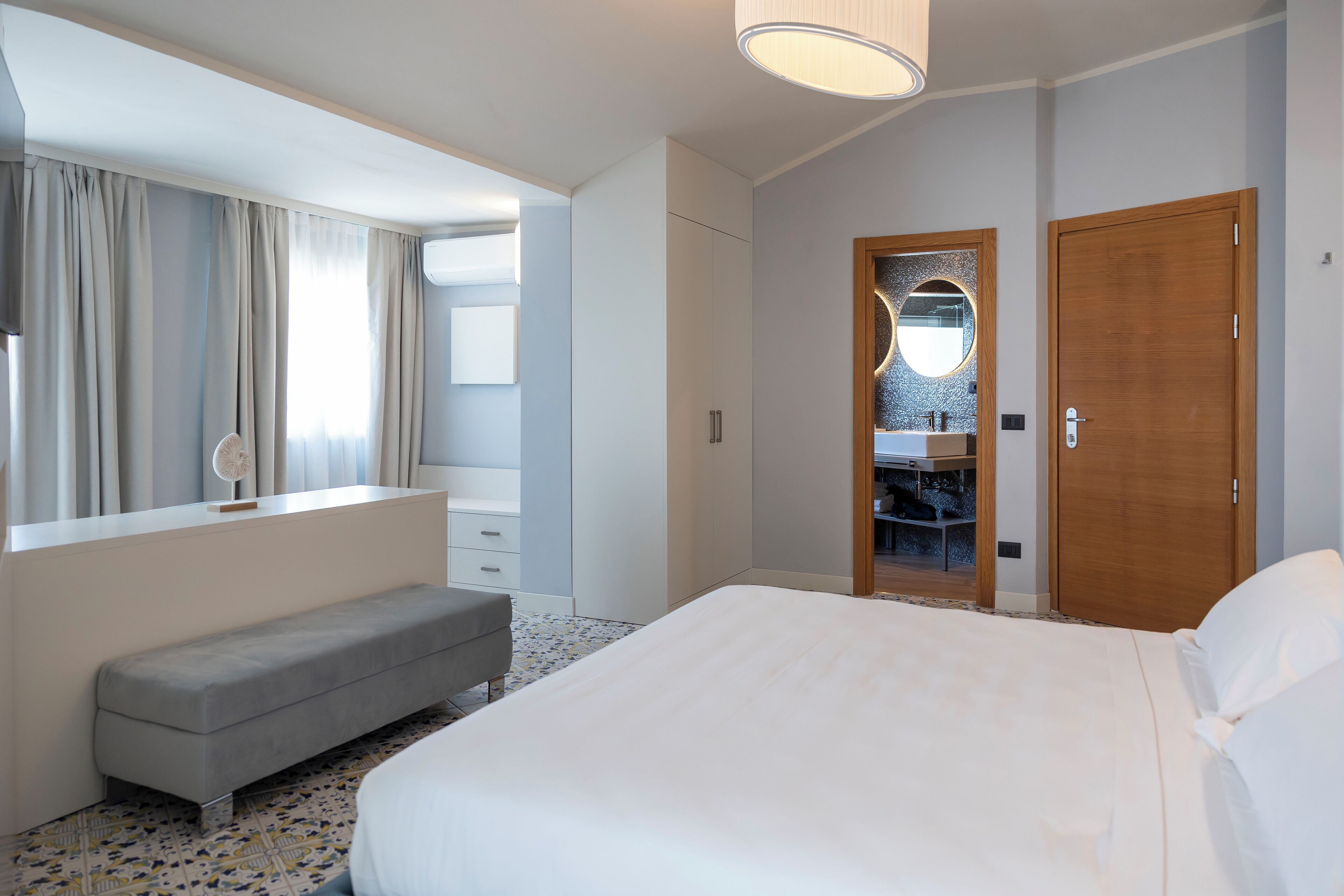 Una camera d'albergo moderna con un letto grande, un comodino, una panca, un armadio e un bagno con specchio e lavandino.