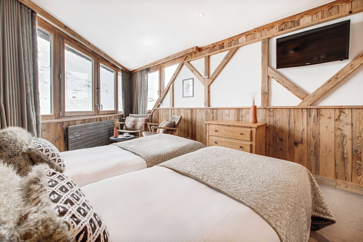 Une chambre d’hôtel avec deux lits, des murs en bois, un téléviseur fixé au mur et une vue sur les montagnes par de grandes fenêtres.